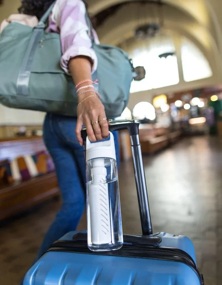 A person standing in a train station holding a transparent water bottle connected to a filtration device inside their suitcase. The person is wearing a pink and white shirt with a large teal shoulder bag, and has a stack of bracelets on their wrist.