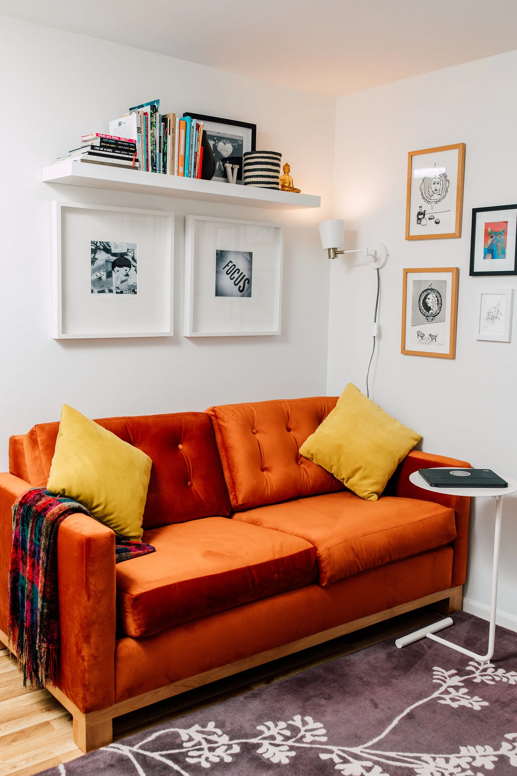 A cozy living room featuring an orange velvet sofa with yellow pillows, a small white side table with a black laptop, and a gray rug with white floral patterns. The wall above the sofa displays framed artwork and a wall-mounted reading light. A white floating shelf holds books, picture frames, decorative objects, and a small statue.