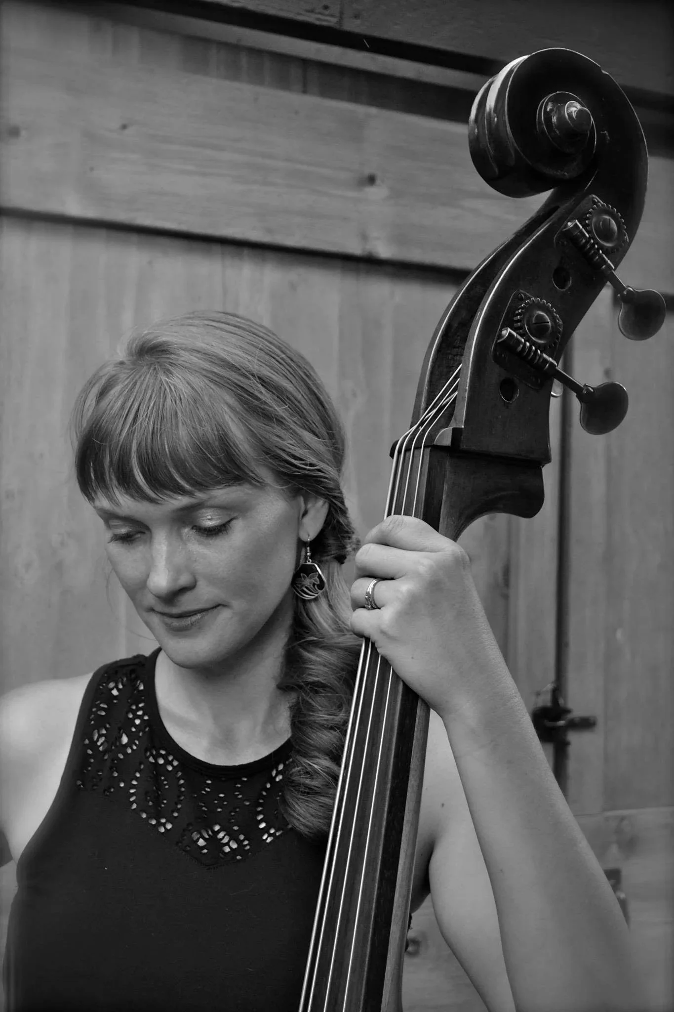 Black and white photo of a woman with closed eyes playing a double bass, with part of her hair and face visible, against a background of wooden panels.