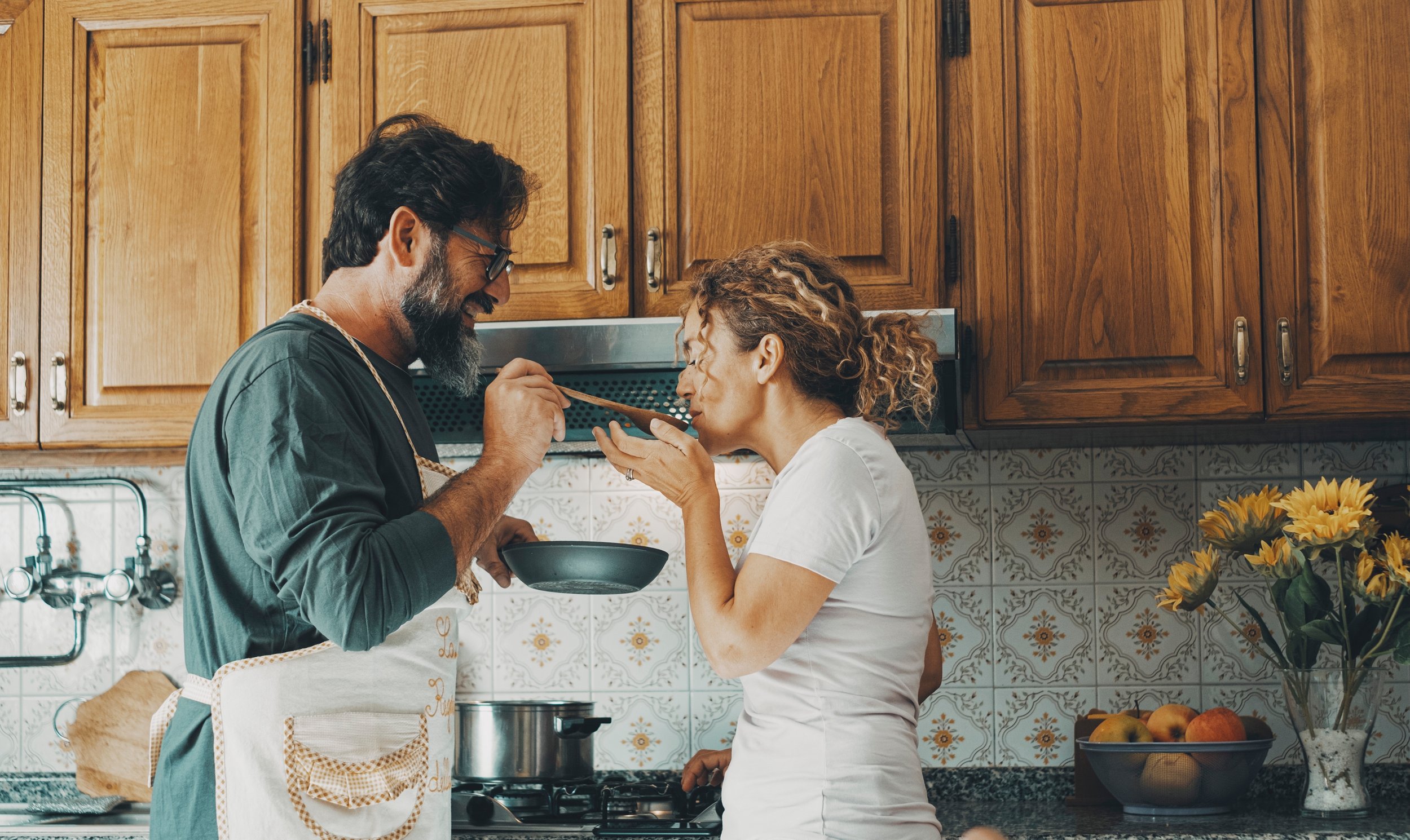 A man and woman in a kitchen, with the man feeding the woman with a spoon.