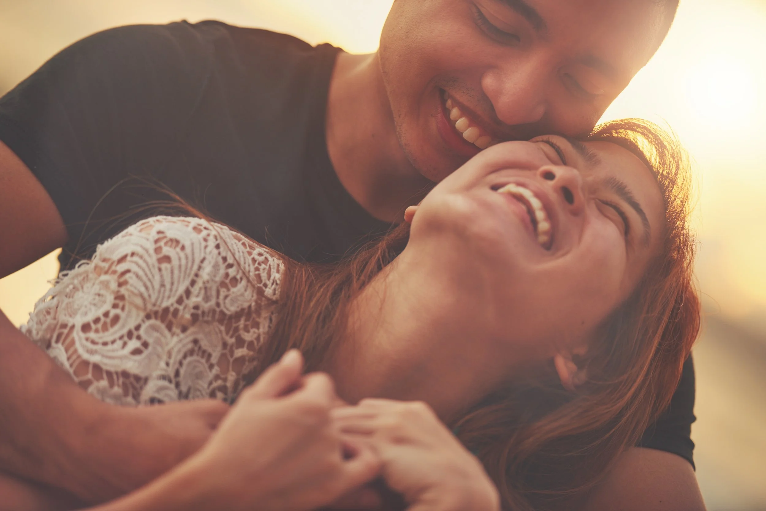 A young man and woman share a joyful, intimate moment, smiling and laughing together in warm, soft lighting.
