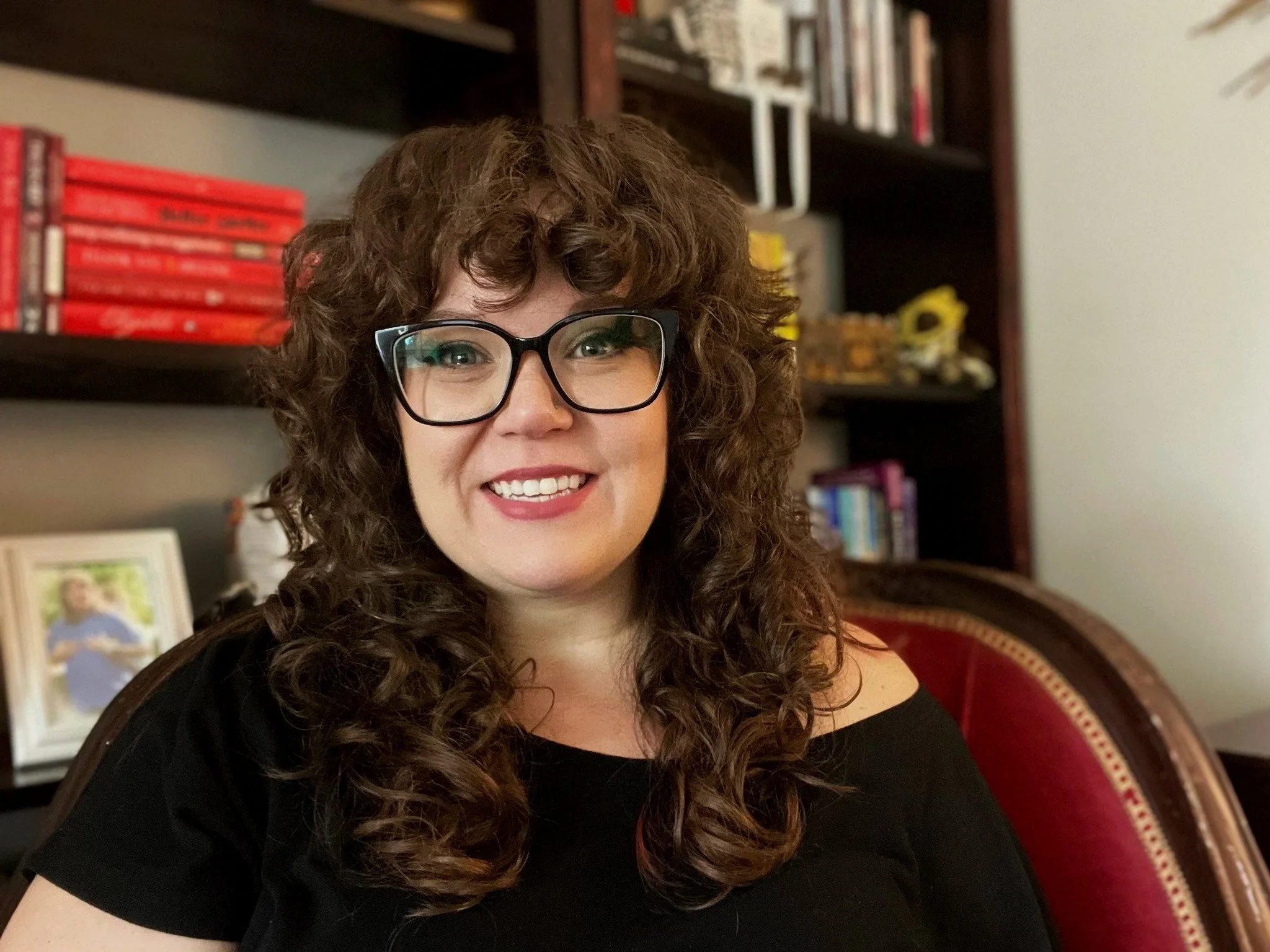 A woman with long curly brown hair, glasses, and wearing a black top, sitting on a vintage red and wood chair, with a bookshelf filled with books and a framed photo in the background.
