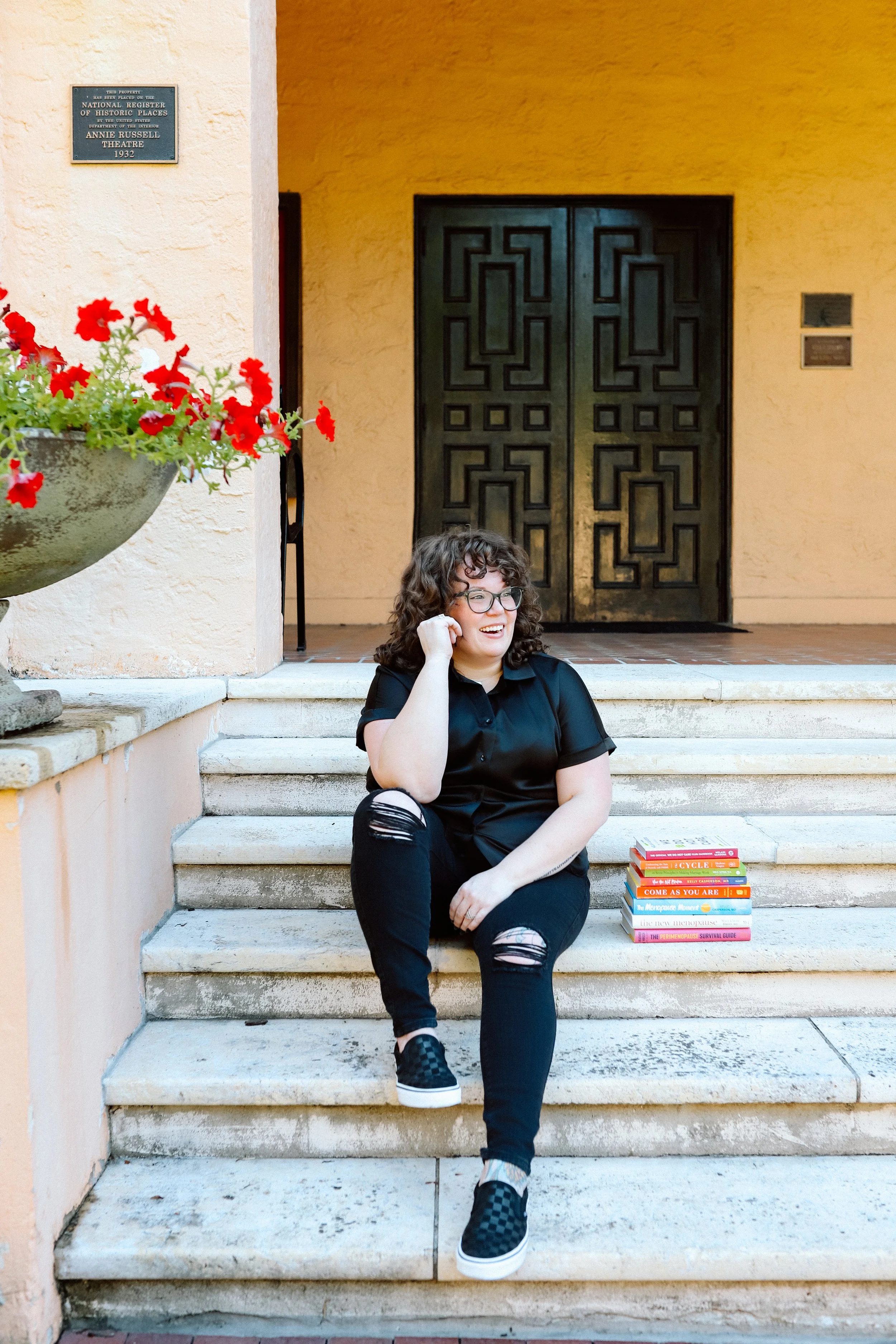 Therapist Mariah Plociniak sitting on outdoor steps beside a stack of relationship and women's health books.