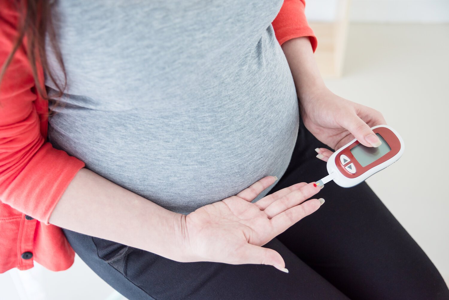 A pregnant woman sitting with her hand on her belly, holding a blood glucose meter, preparing to check her blood sugar level.