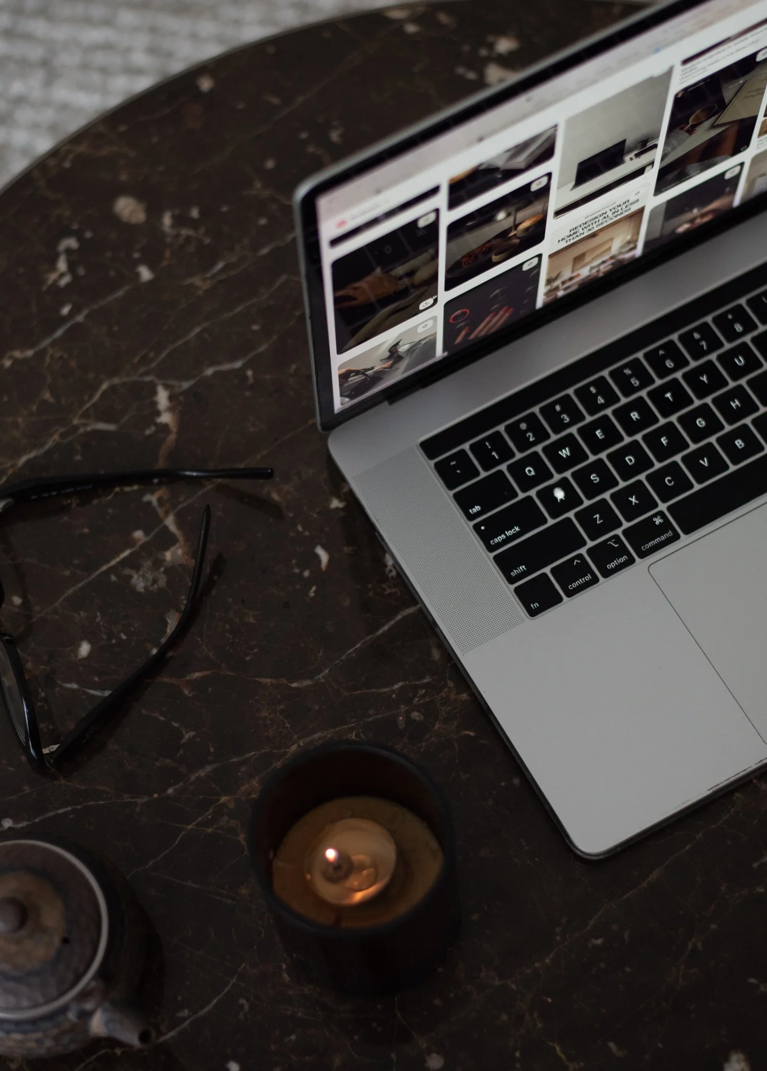 A laptop on a dark marble table displaying a grid of images, with a pair of black eyeglasses and two lit candles nearby.