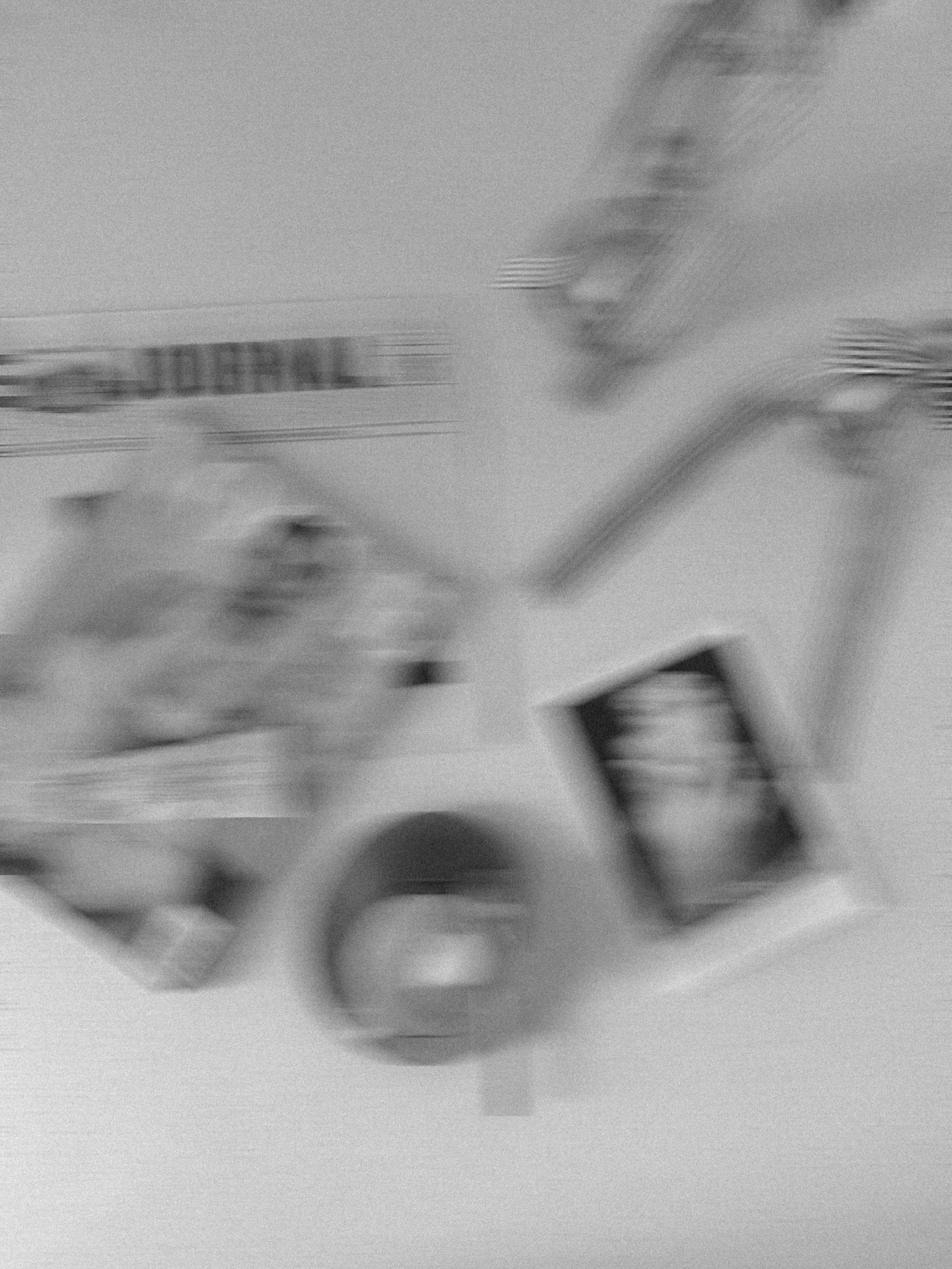 Black and white blurred photo of various objects including a cup, a photograph, and some boxes or containers, with some text in the background.