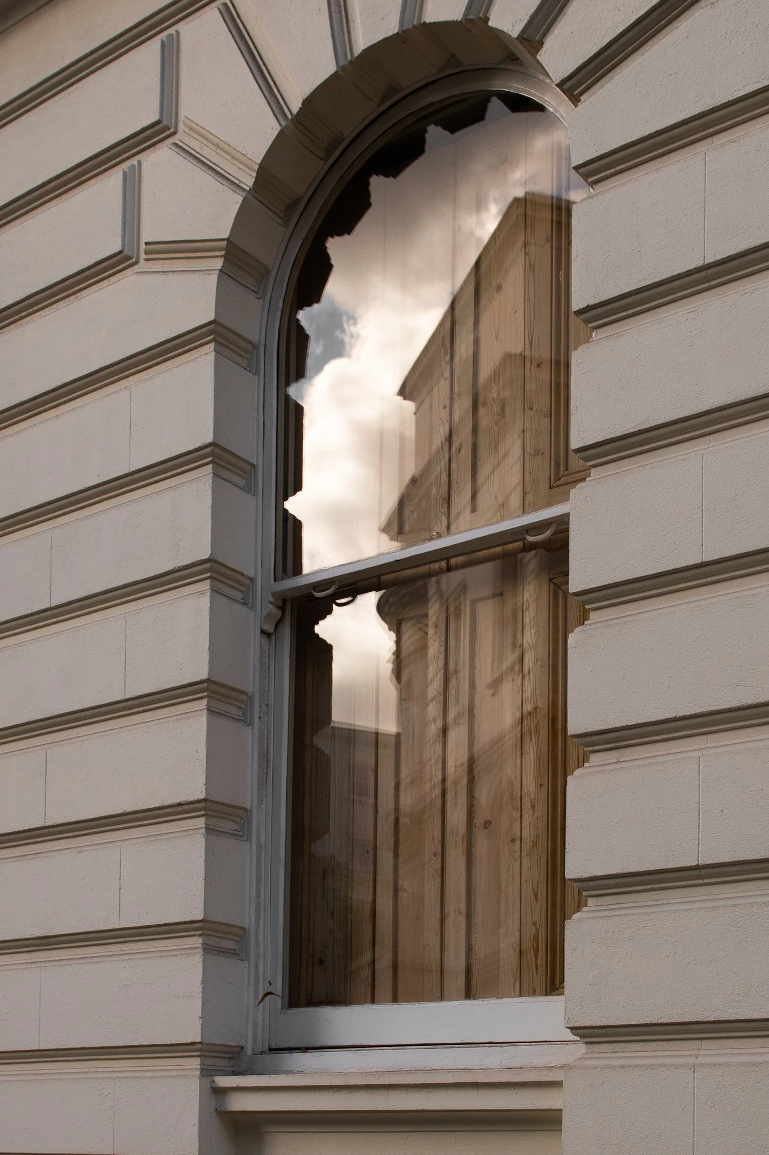 Broken window on a cream-colored building, with a view of the sky and a wooden structure reflected in the glass.