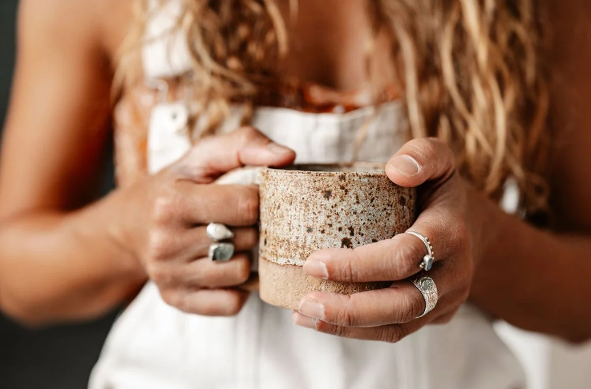 Close-up of a person holding a beige, speckled ceramic mug with both hands, wearing rings and a white apron, with long, curled hair visible.
