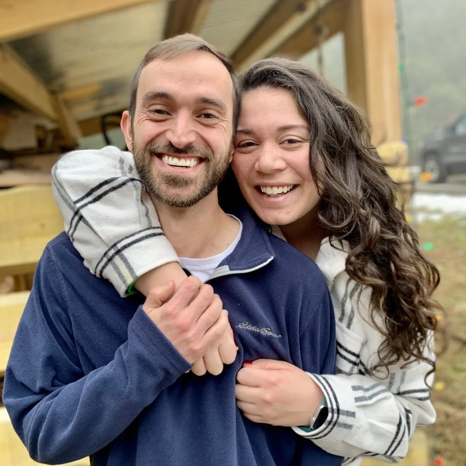 A husband and wife are smiling for a portrait, with her arms wrapped around him.