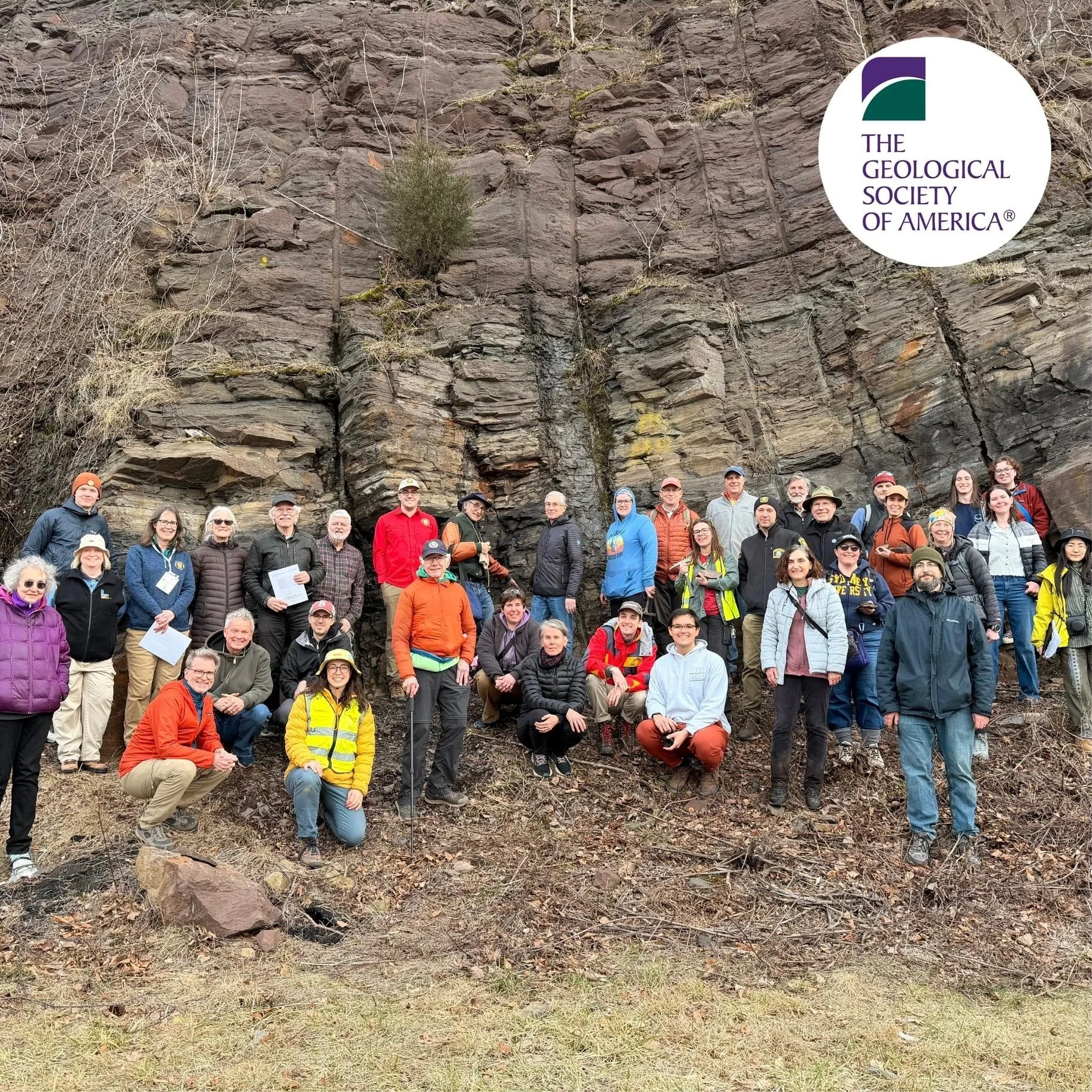 GSA Field Trip participants in front of a tall, rocky cliff with sparse vegetation. Some are standing, and some are sitting on the ground, facing the camera. Many are smiling, dressed in casual outdoor clothing, with some wearing hats and jackets.