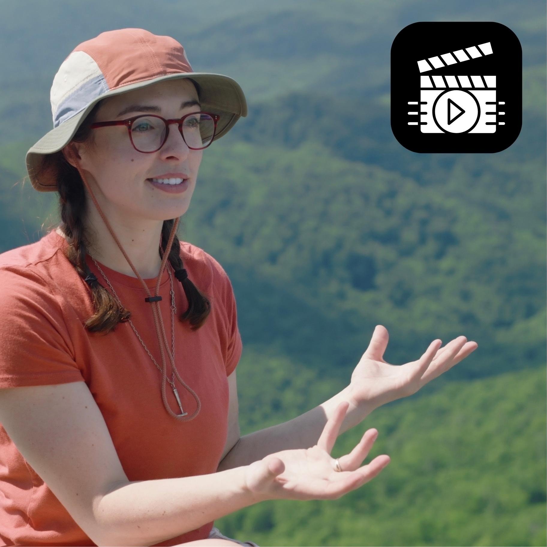 Close up of Geo Girl wearing glasses and a layered sun hat is outdoors with a blurred green landscape in the background. She is wearing a red shirt and has her hands raised, possibly explaining or gesturing.