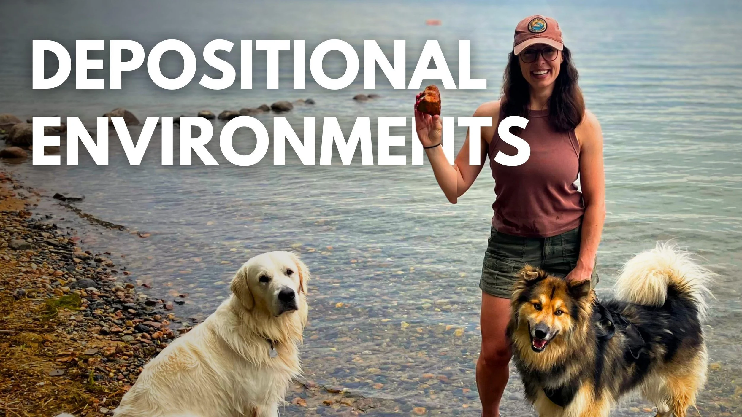 Geo Girl with glasses and a pink cap standing by a lake with two dogs, holding an agate from the shore. The lake has a rocky shoreline and calm water with a few floating objects.