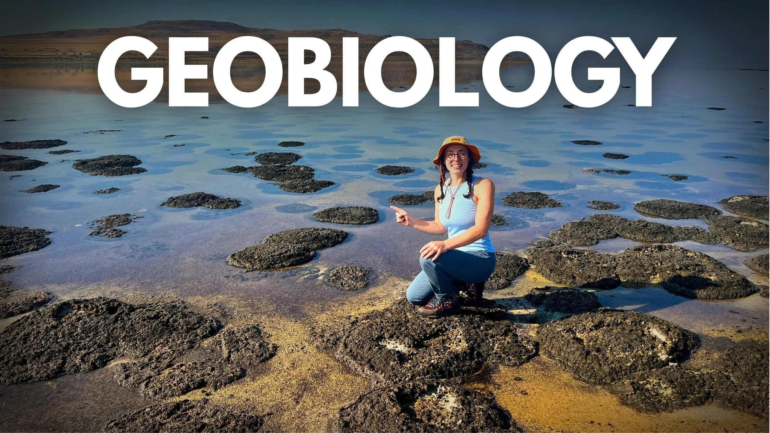 Geo Girl kneeling on rocky terrain of stromatolites near a body of water, smiling and wearing a sunhat and glasses, with large white text reading 'GEOLOGY' across the top of the image.