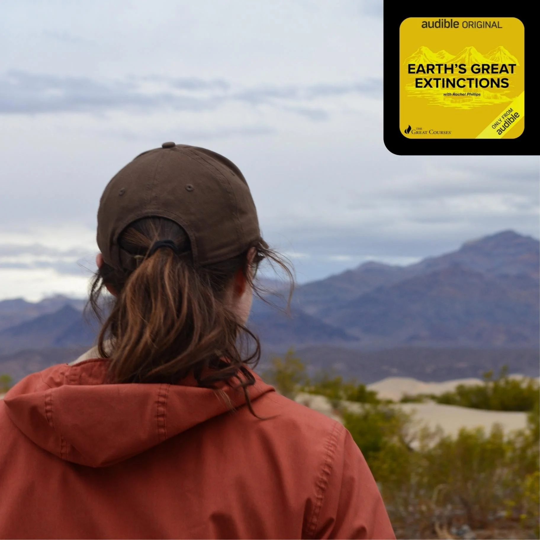 Photo of Geo Girl from behind with reddish-brown hair wearing a brown baseball cap and a reddish jacket, looking at a mountain landscape with cloudy skies in the background.