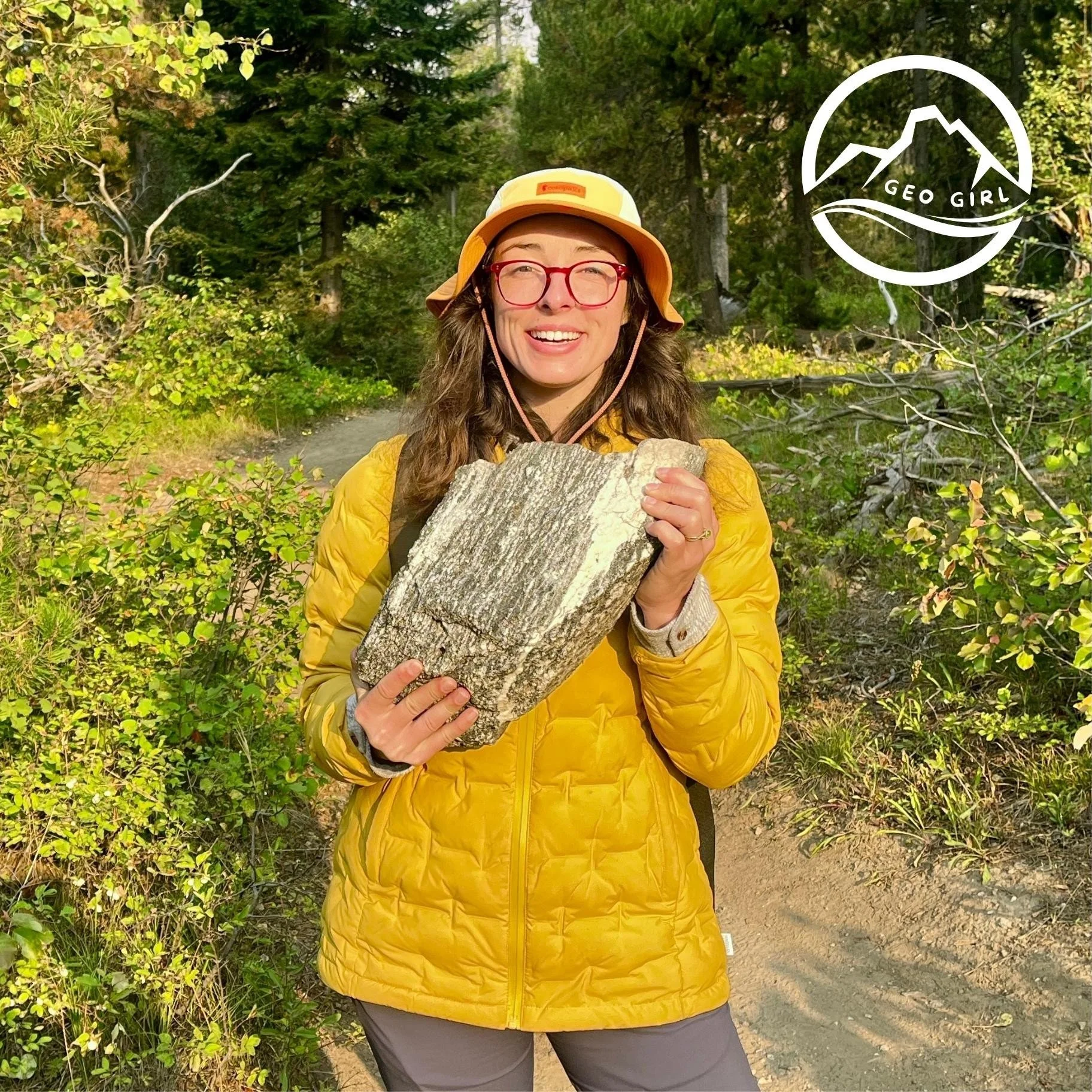 Geo Girl wearing a yellow outdoor jacket, red glasses, and an orange hat holding a piece of gneiss in a lush green forest of Grand Teton National Park.
