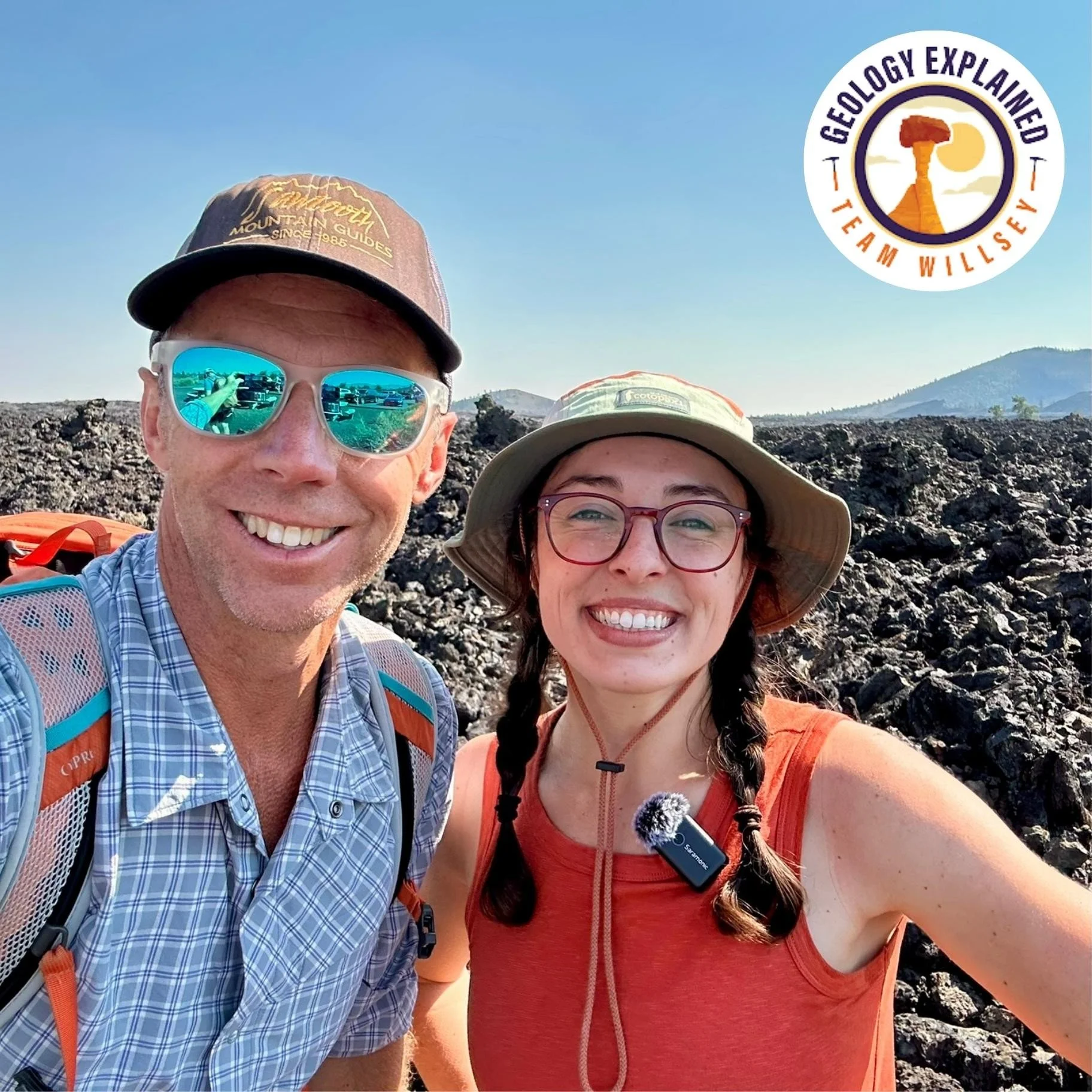 Shawn Willsey and Geo Girl taking a selfie outdoors in a rocky volcanic landscape, with a distant cinder cone and clear blue sky in the background. 