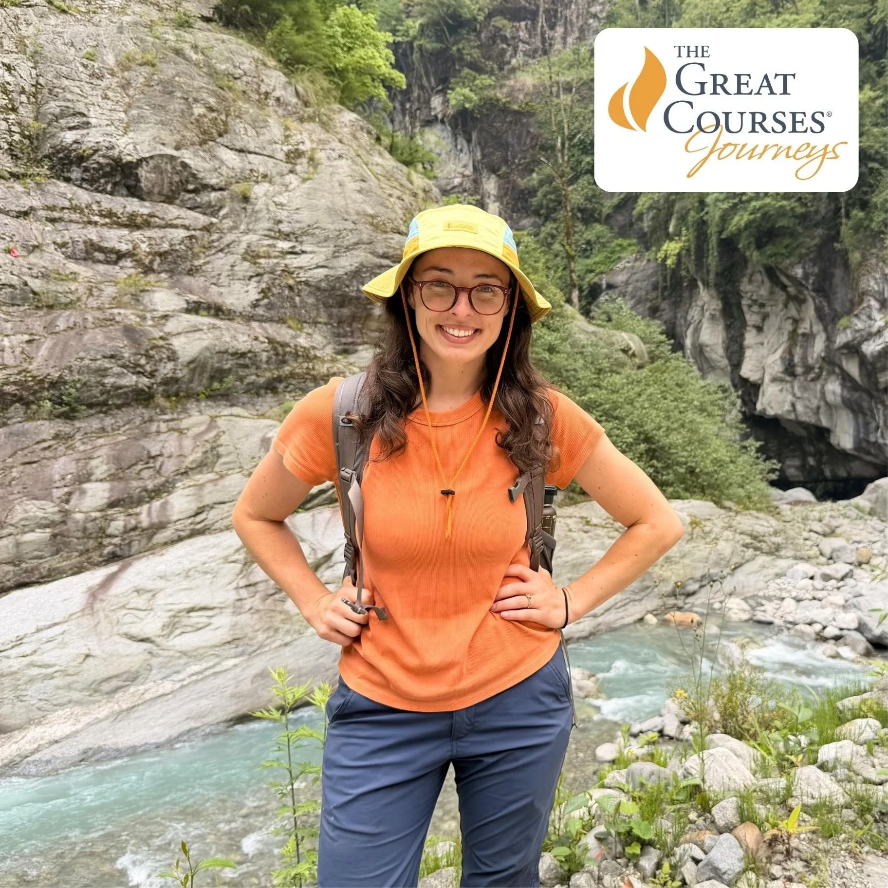 Geo Girl smiling and posing outdoors near a river with rocky cliffs and green trees in the background. She is wearing a yellow hat, orange shirt, and glasses, with a backpack on her shoulders.