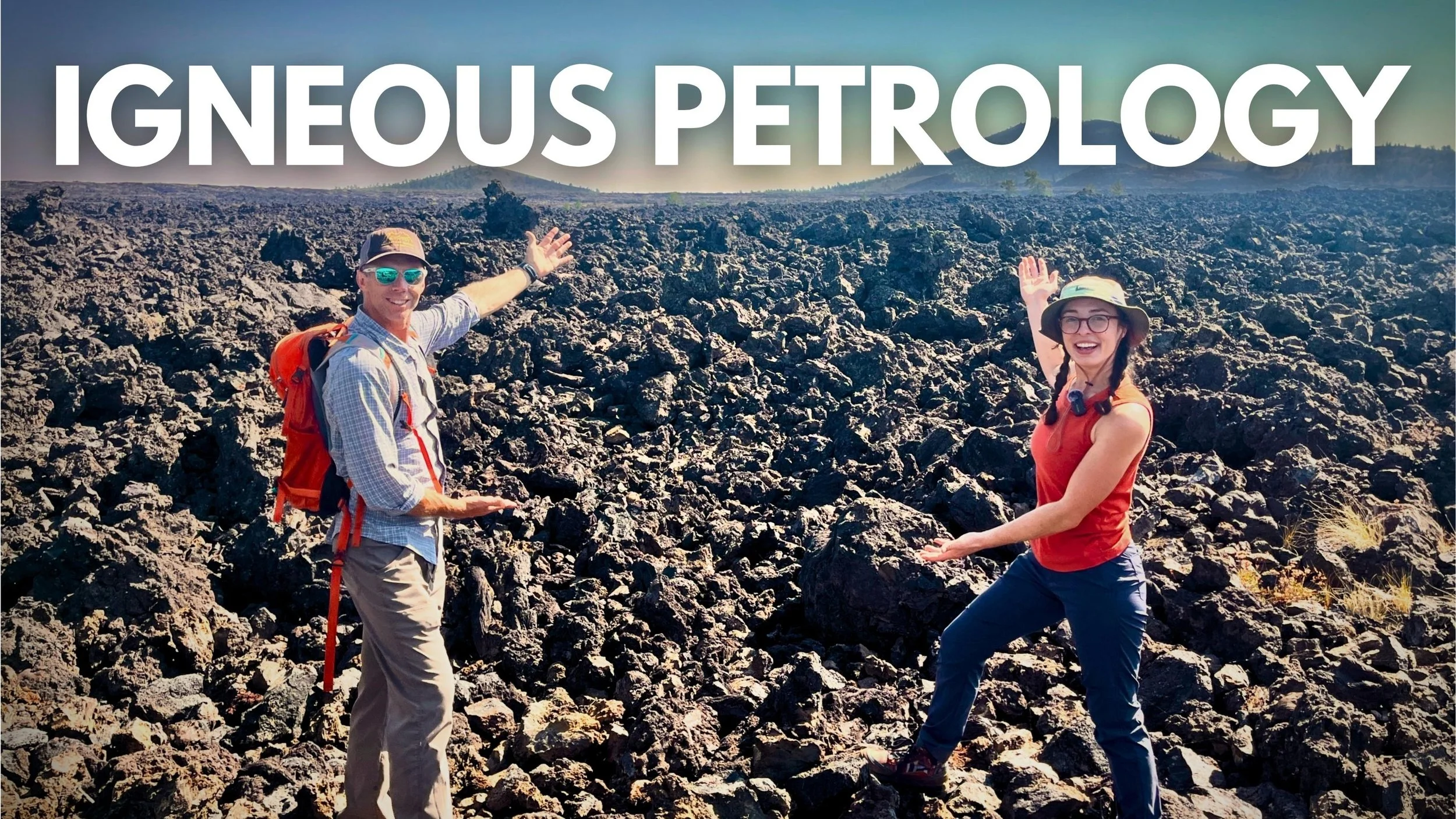 Geo Girl and Shawn Willsey standing on a rocky basaltic lava flow volcanic landscape, pointing and smiling at the camera, with distant hills and a blue sky in the background.