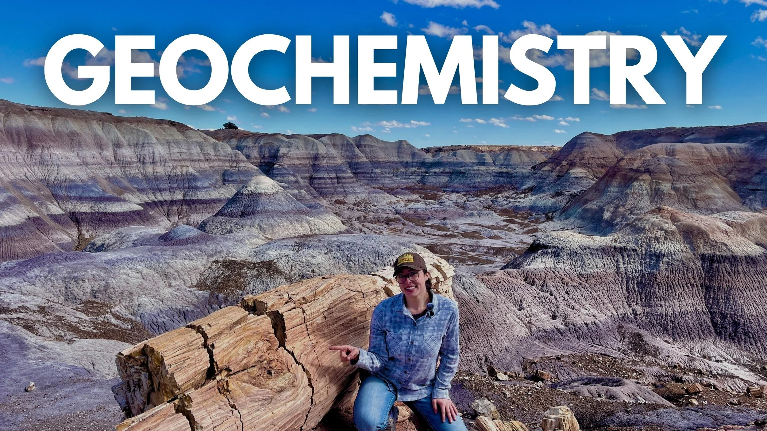 Geo Girl wearing glasses and a plaid shirt, pointing at a large piece of petrified wood in a desert landscape with colorful layered rock formations and a blue sky with clouds behind her.
