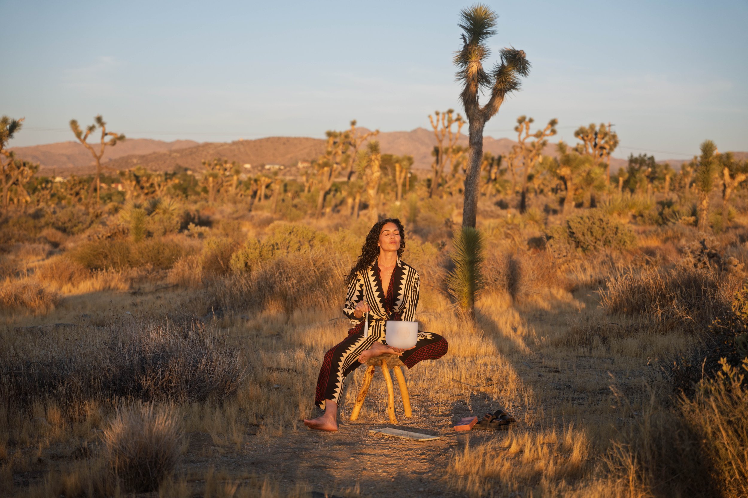 A person with long dark hair practicing yoga on the beach, with arms raised above head and hands pressed together in a prayer position, facing the ocean.