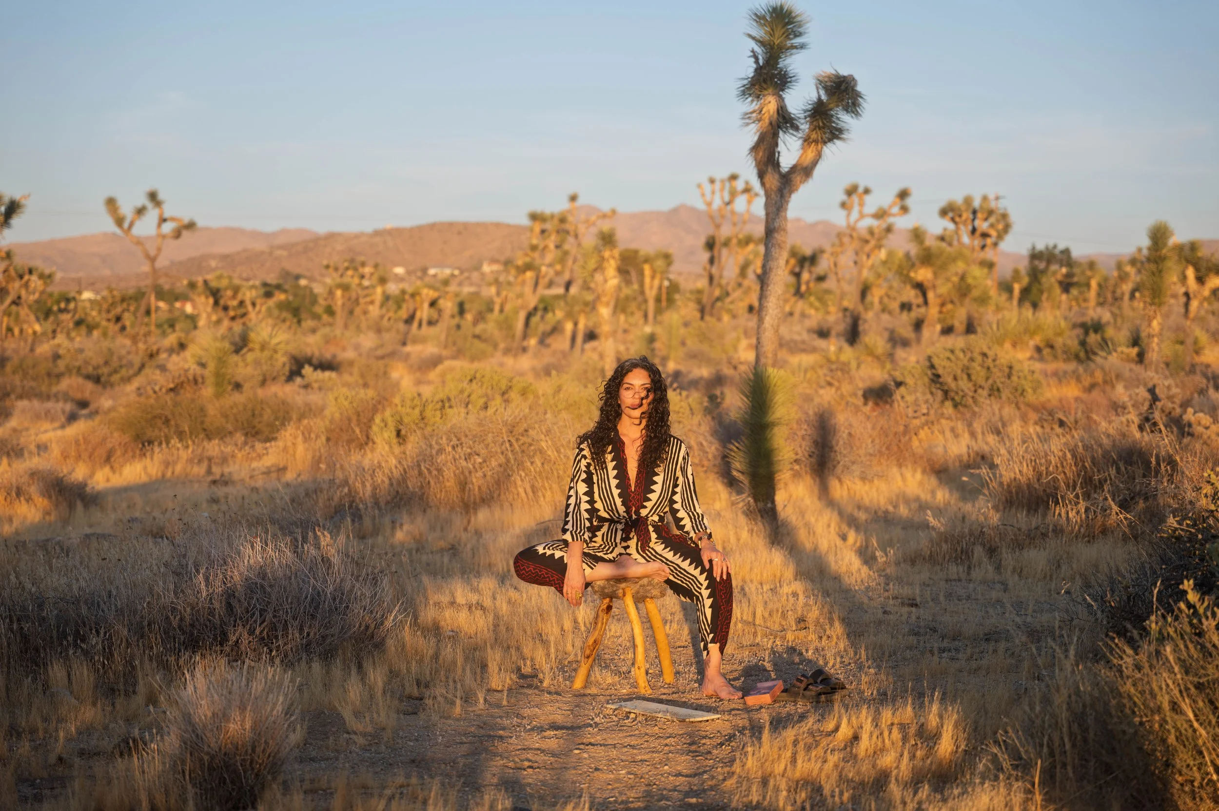 A woman with long, wavy hair practicing yoga outdoors by the ocean, wearing a blue sports bra and matching leggings, executing a balancing pose with her right leg extended and her right arm reaching over her head.