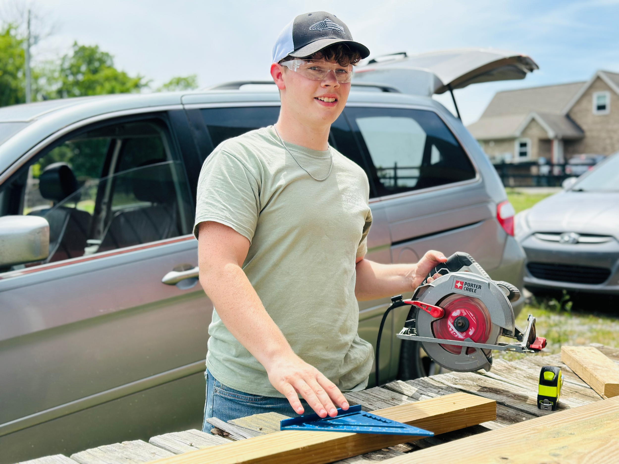 A young man wearing safety glasses, a black and white cap, and a light green t-shirt is smiling while working outdoors on a woodworking project, holding a circular saw, with a car and houses in the background.