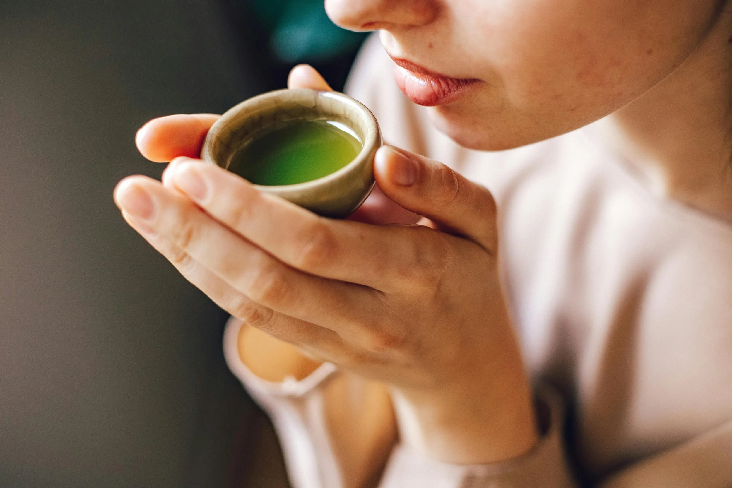 A woman holding a small cup of green tea close to her lips, preparing to drink.