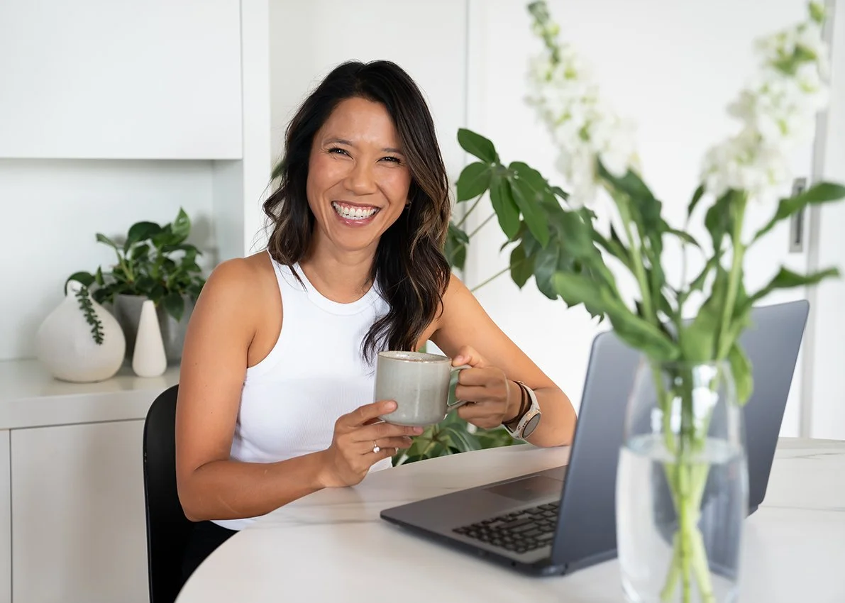 Woman smiling and holding a mug at a desk with a laptop and green plants in a bright, modern room.