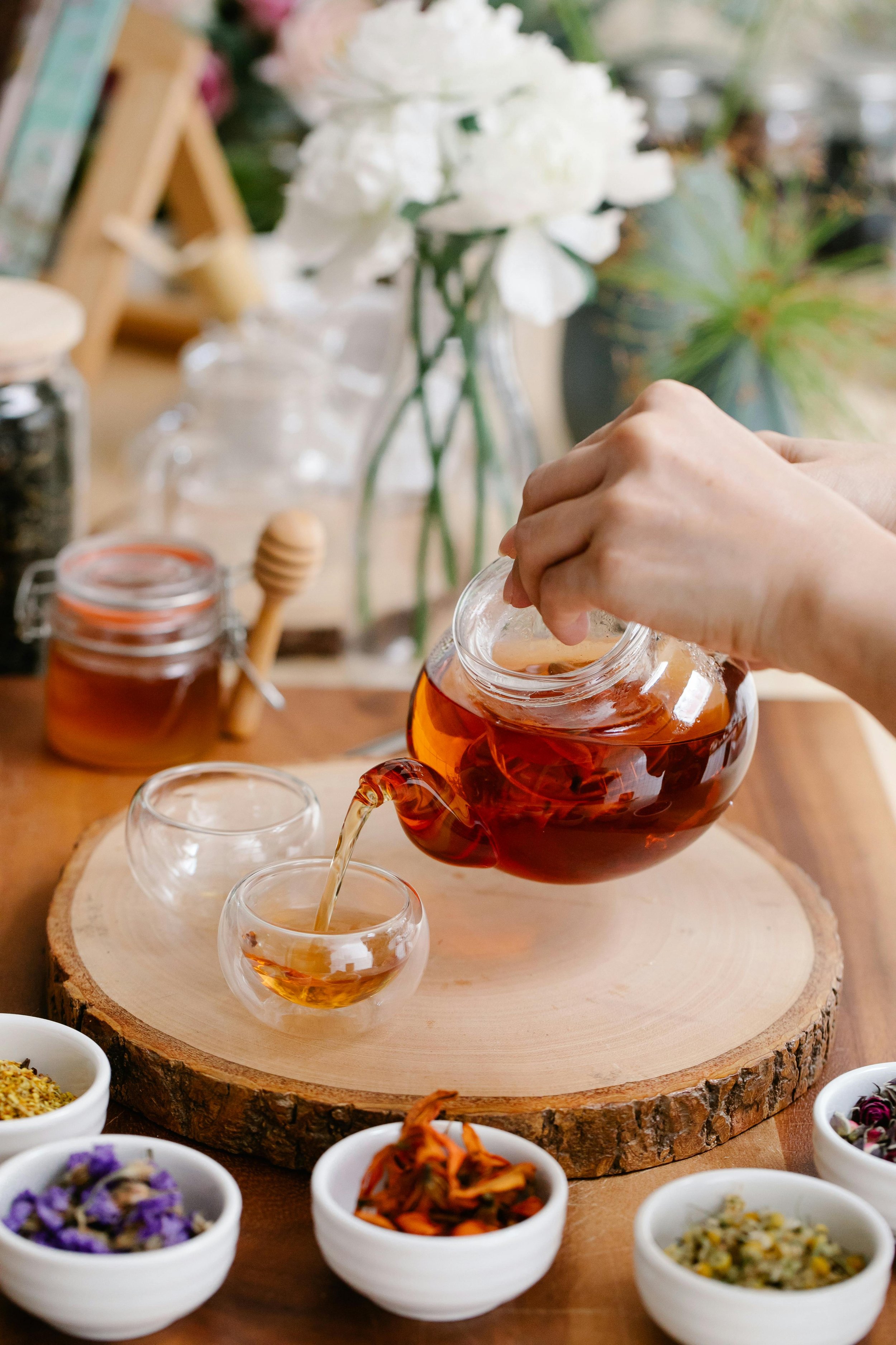 Person pouring tea from a glass teapot into small glass cups on a wooden surface with bowls of dried herbs and flowers in the foreground and a vase of white flowers in the background.