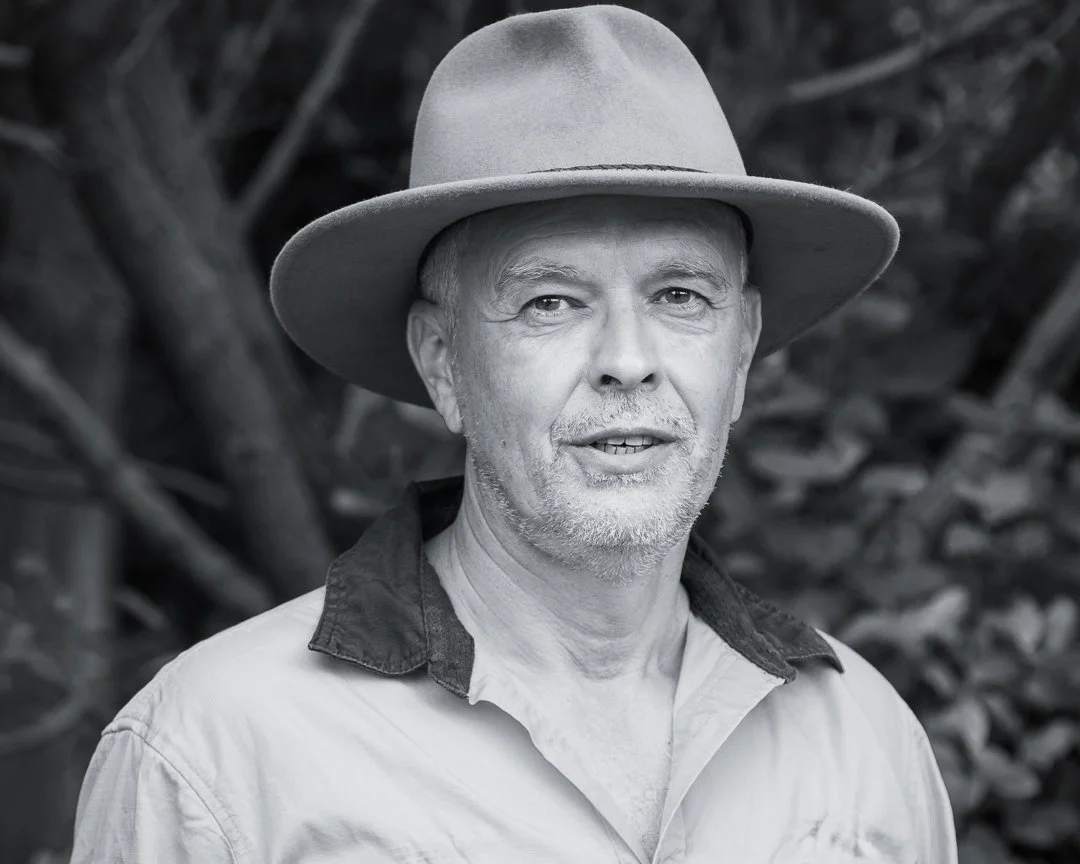 A black and white portrait of a middle-aged man wearing a wide-brimmed hat and a casual shirt, standing outdoors with blurred foliage in the background.