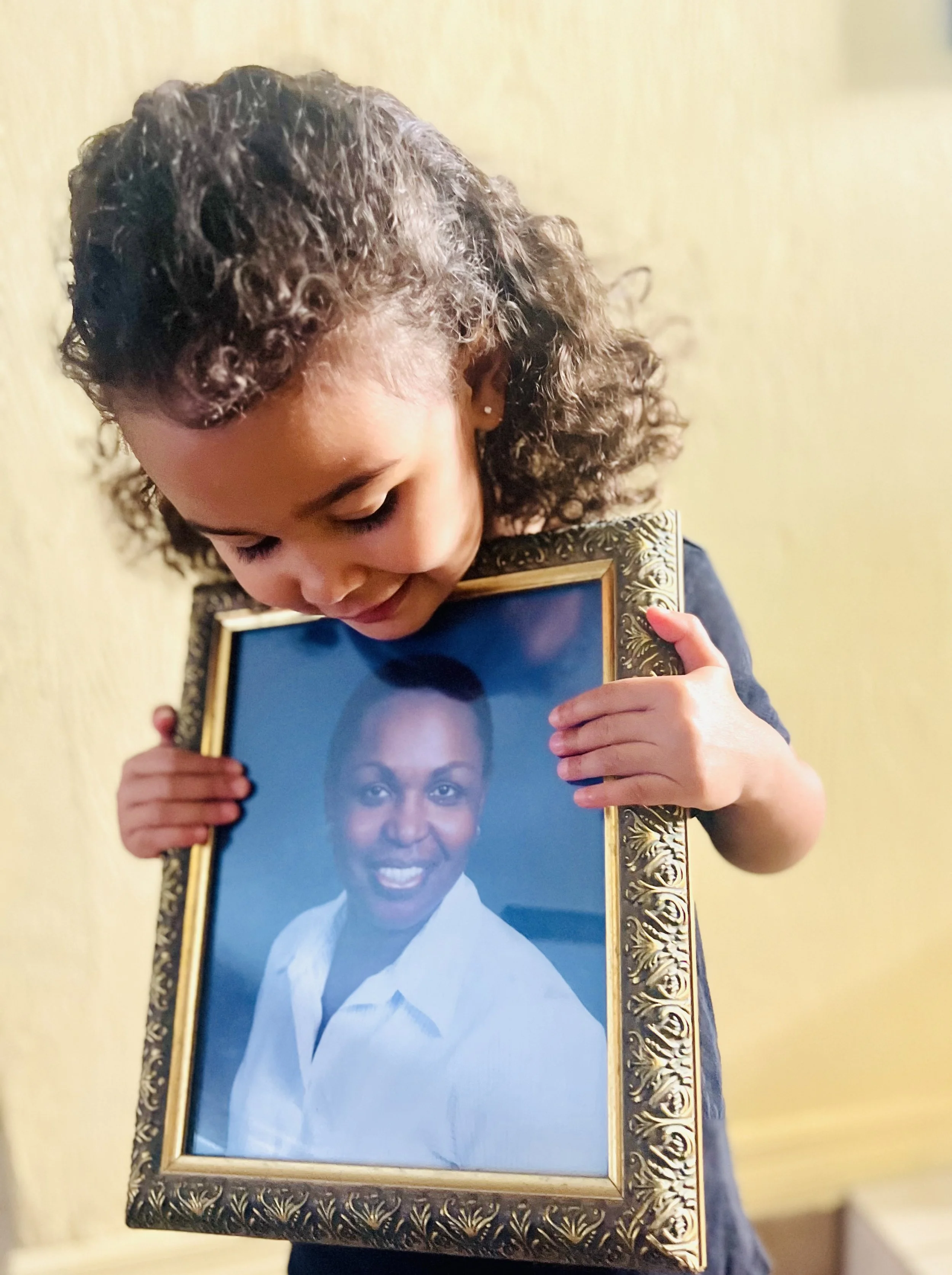 A young girl with curly hair holding a framed portrait of an older woman with short dark hair and a white collared shirt, looking down and smiling.