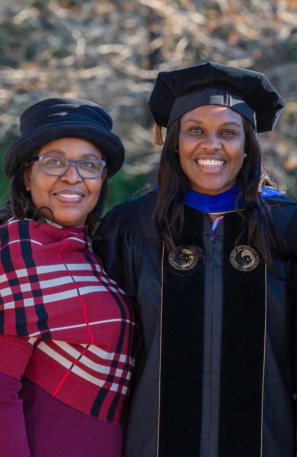 A woman in graduation regalia standing between two adults outdoors on a sunny day, with trees in the background.