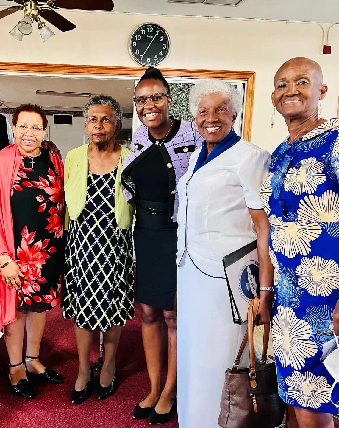 A group of seven diverse people, four women and three men, pose together indoors, smiling for the camera. There is a clock on the wall behind them showing about 7:03, and a ceiling fan is visible.