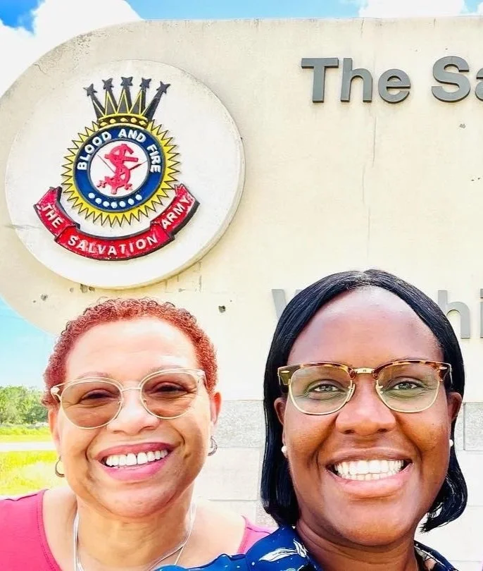 Group of three people smiling in front of 'The Salvation Army' Center sign, with the Salvation Army emblem visible, during daytime.
