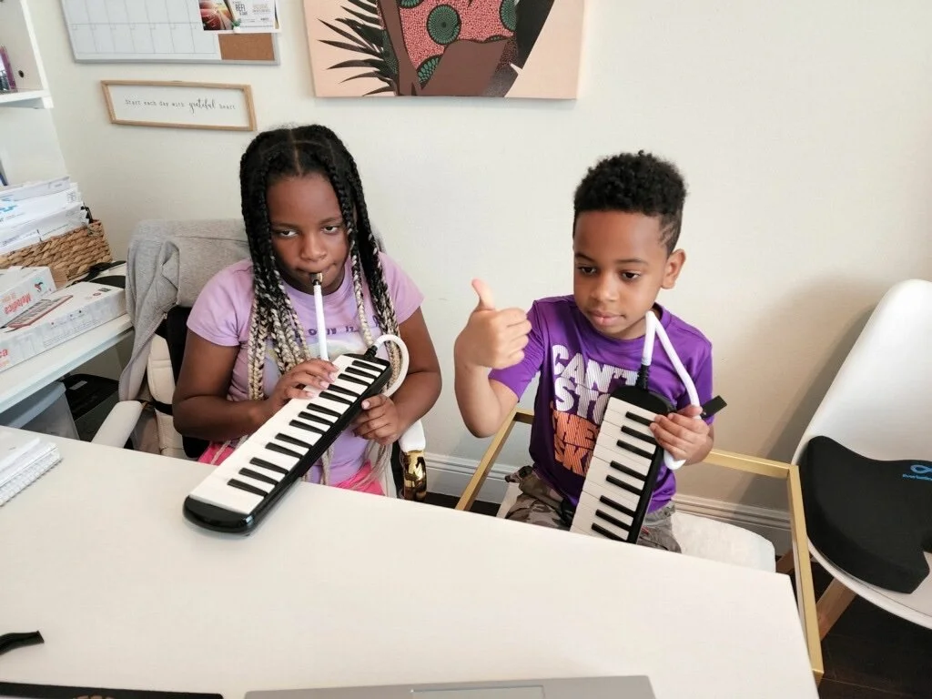 Two young children sitting at a desk, each holding a toy keyboard and making playful expressions. The girl on the left has braided hair and is pretending to play the keyboard, while the boy on the right has short curly hair and is giving a thumbs-up.