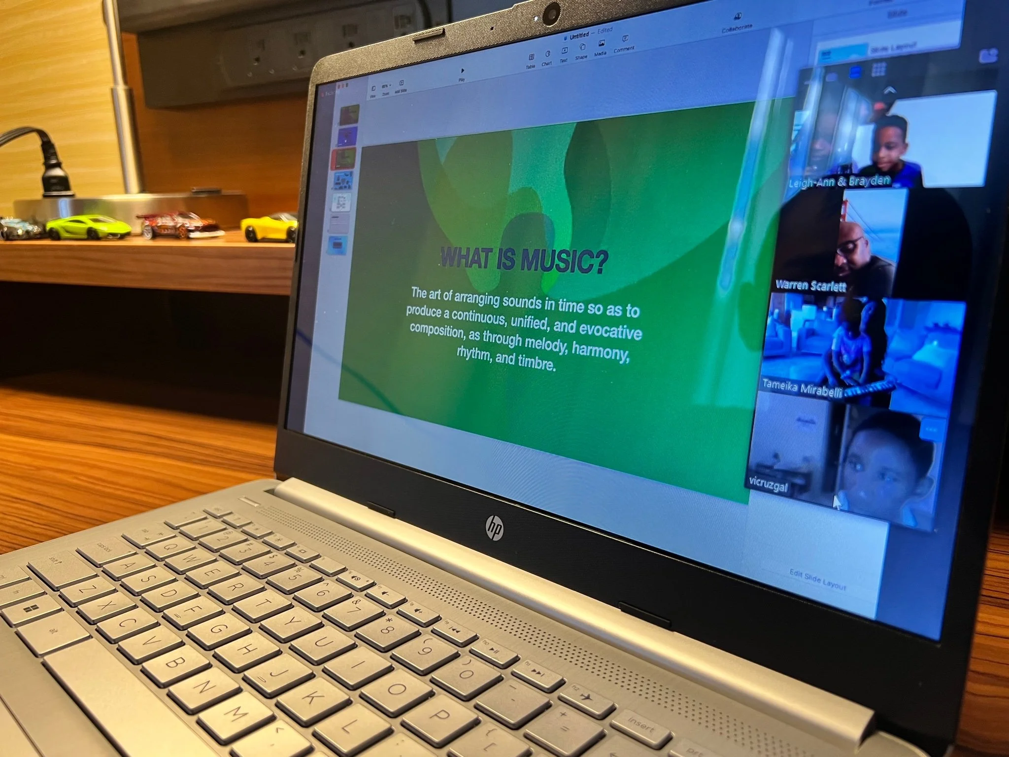 A laptop on a wooden table displays a presentation slide titled 'What is Music?' with a green background, explaining that music is arranging sounds in time to produce harmony, melody, rhythm, and timbre. To the right, a Zoom video call shows four chi
