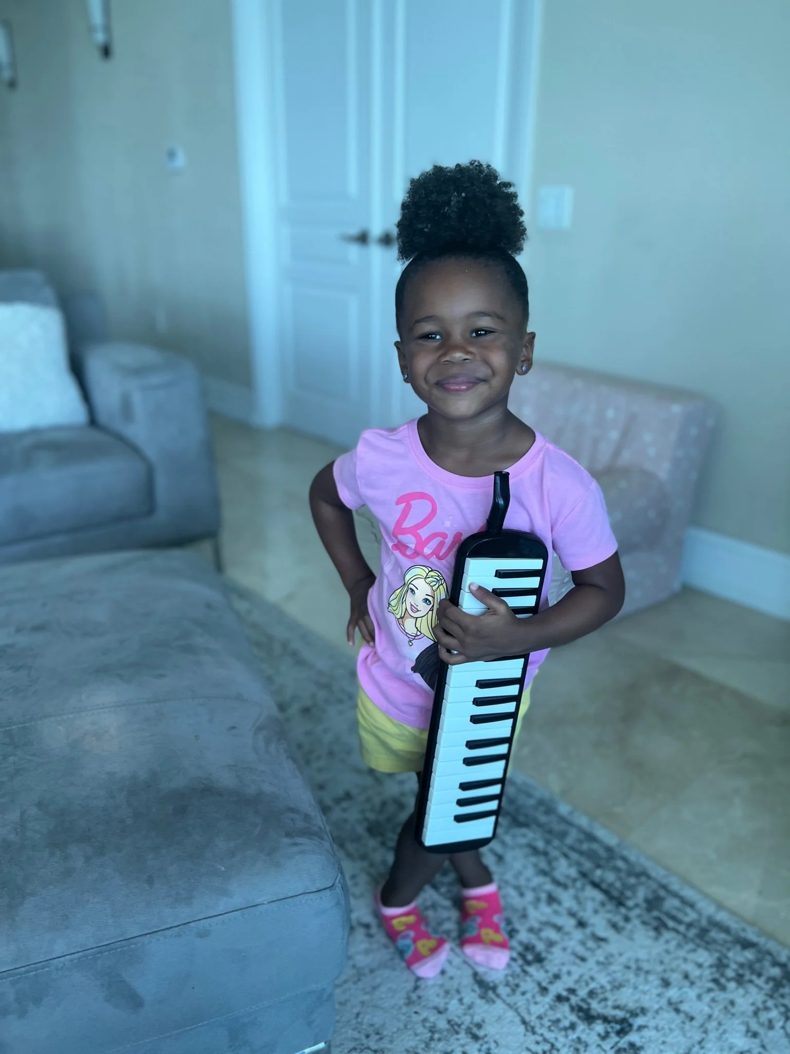 A smiling young girl with curly hair in a high puff, wearing a pink Barbie t-shirt and yellow shorts, holding a mini keyboard toy.