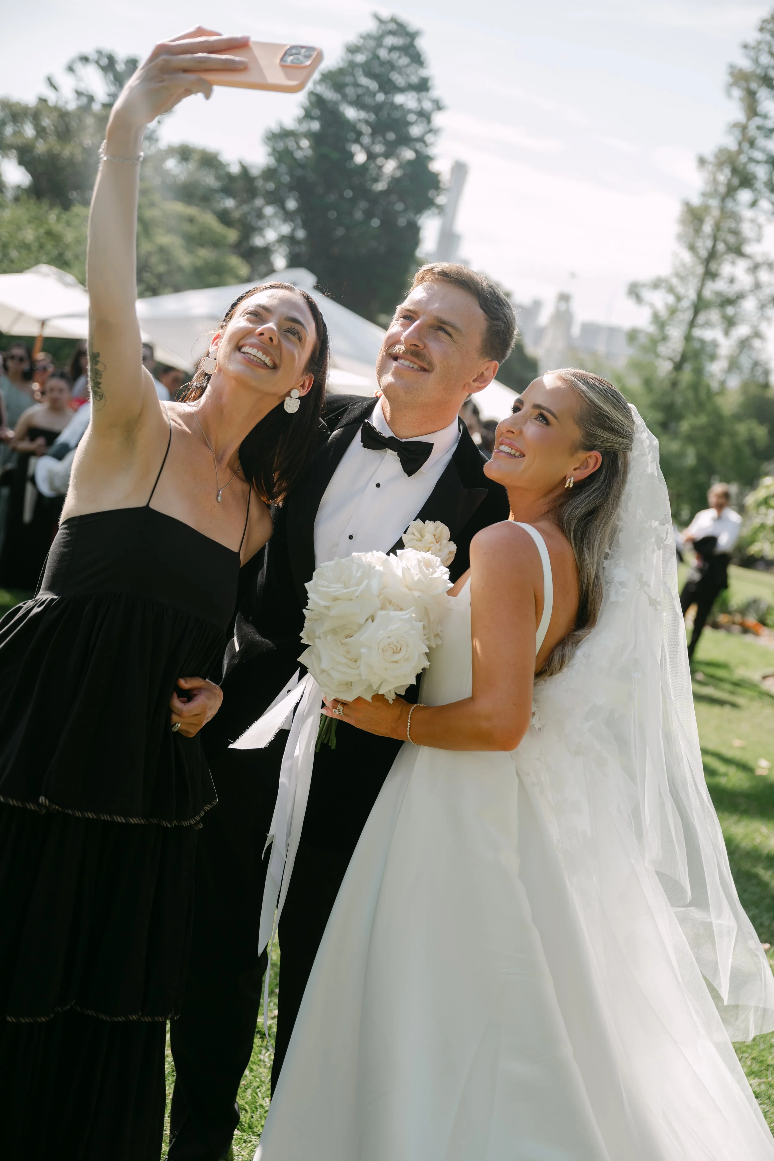 A woman in a black dress taking a selfie with a man in a tuxedo and a woman in a white wedding dress holding a bouquet at an outdoor wedding reception.
