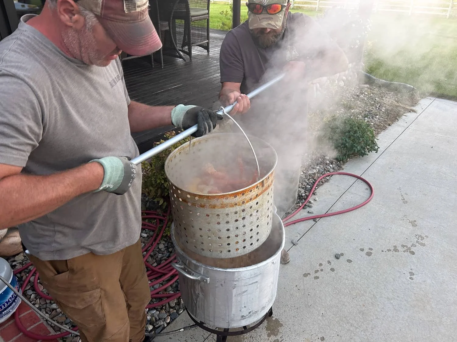 Two men outdoors, one wearing a gray shirt, tan shorts, a cap, and gloves, lifting a steaming metal pot with a long-handled pole from a large metal barrel, while the other man, with a beard, sunglasses, hat, and black shirt, observes and helps.