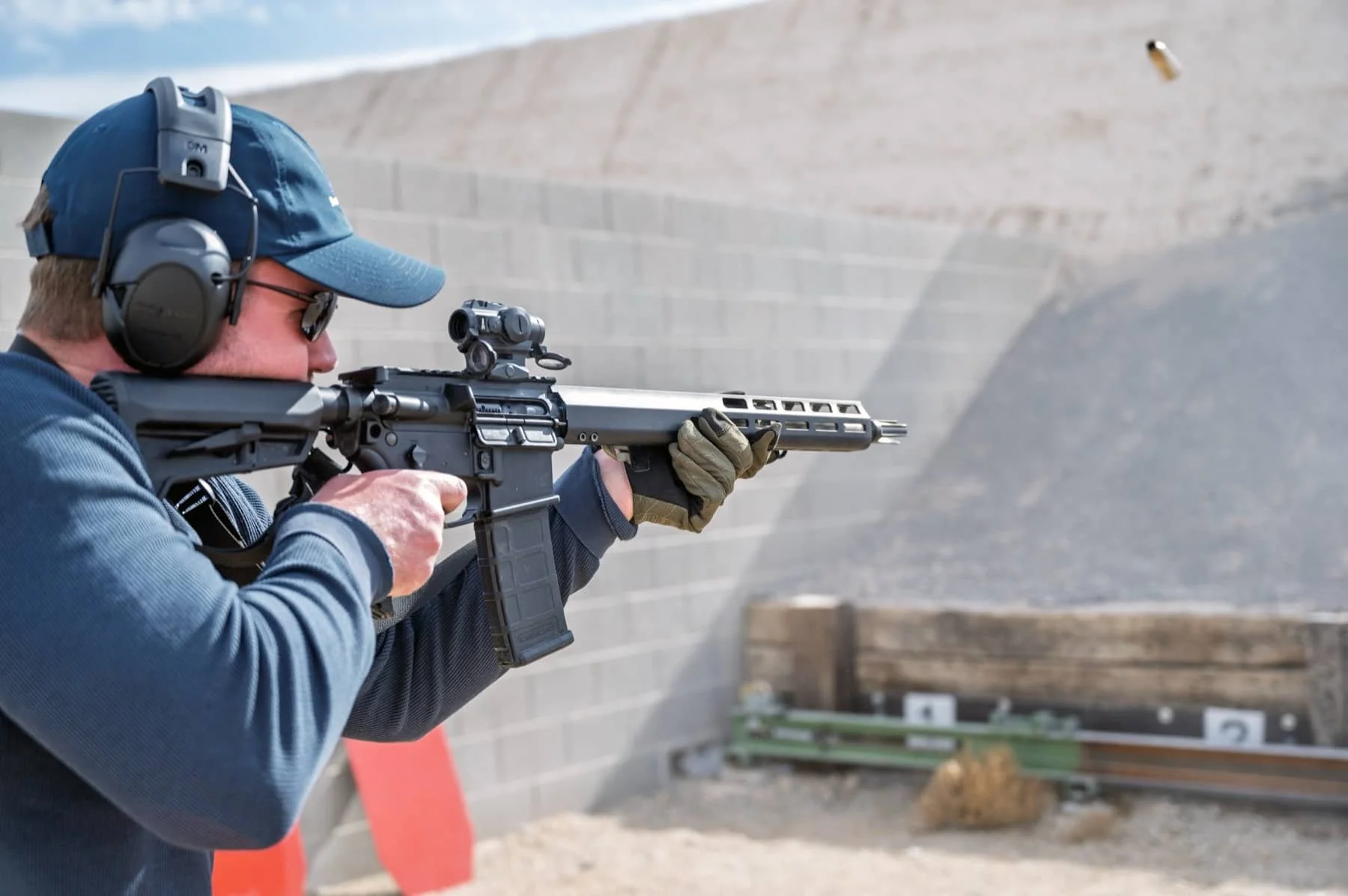 Man at outdoor shooting range aiming rifle with scope, wearing ear protection, sunglasses, and a blue cap.