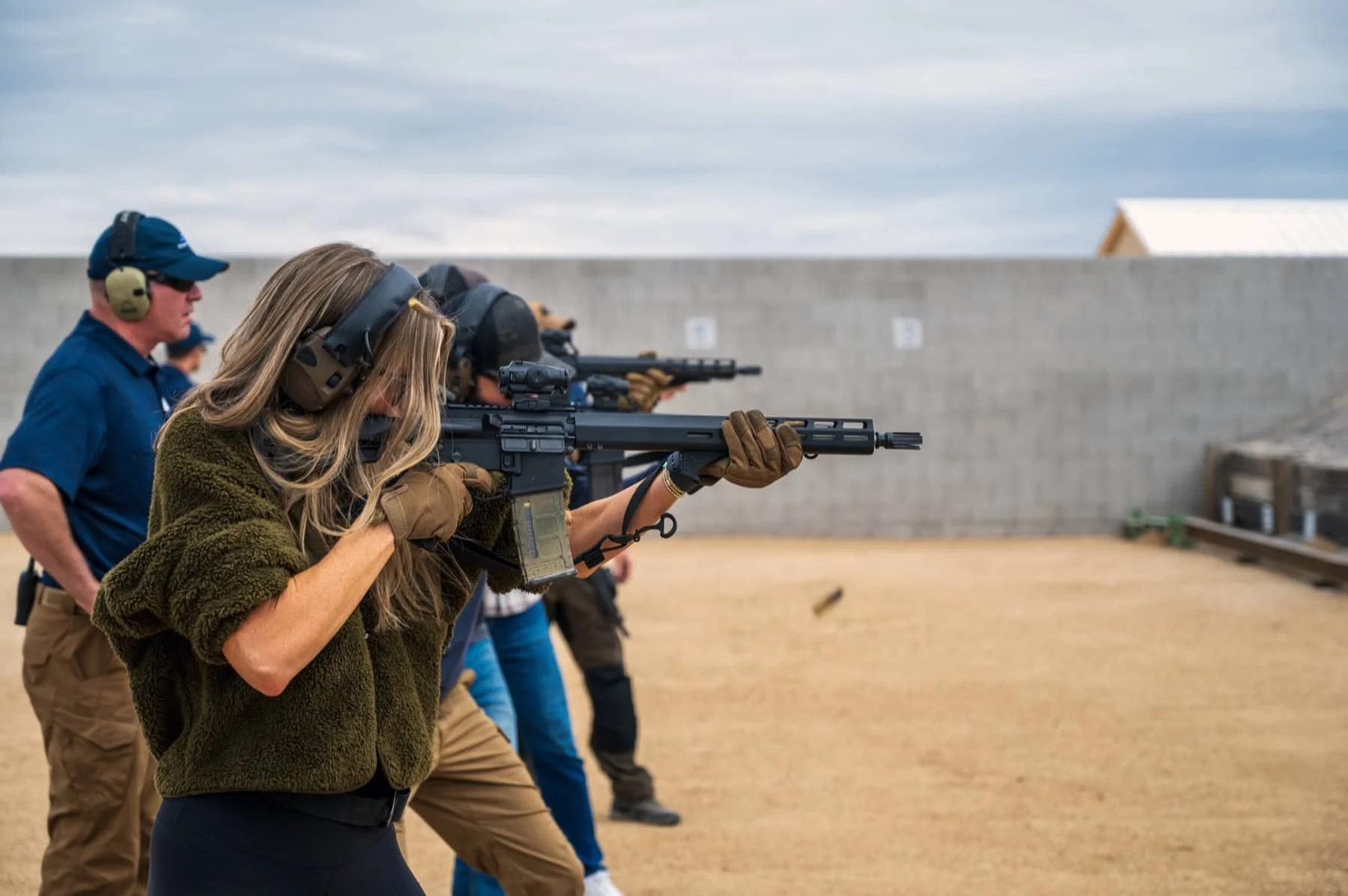 Multiple individuals participating in a shooting practice, aiming rifles at a target range outdoors, with a concrete wall and cloudy sky in the background.