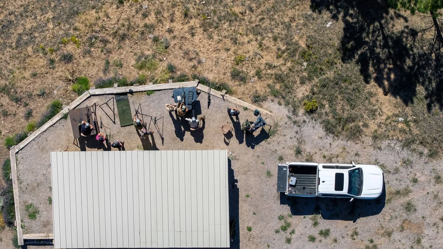 An aerial view of a small outdoor gym with exercise equipment, a group of people working out, and a white pickup truck parked nearby, next to a building with a metal roof, surrounded by a gravel and dirt area with sparse vegetation.