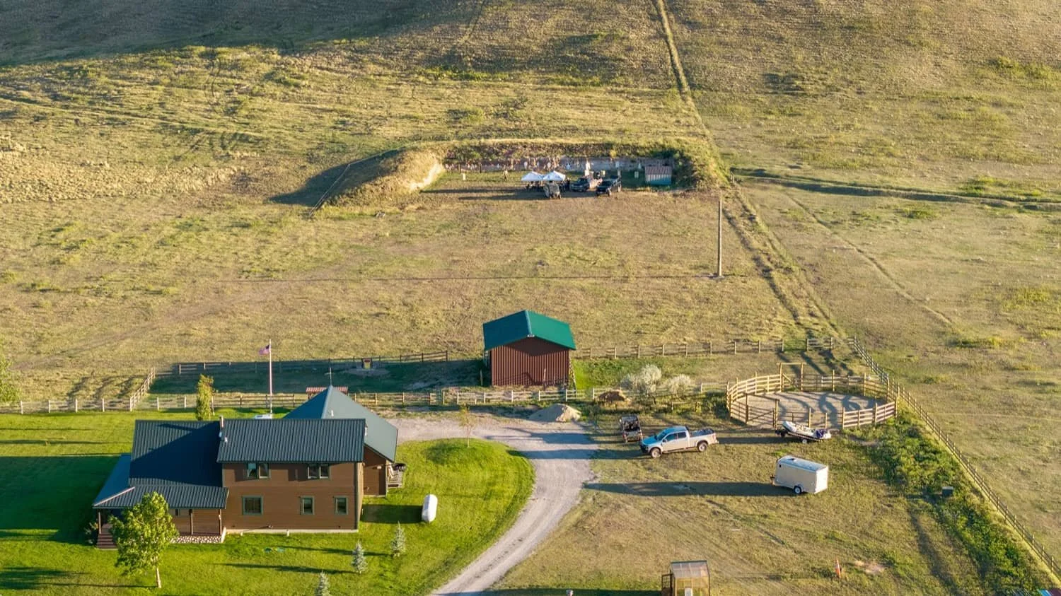 Aerial view of a rural property with a brown house, green lawn, garden, fence, driveway, several vehicles, a small red barn, and vast farmland with some structures and trees.