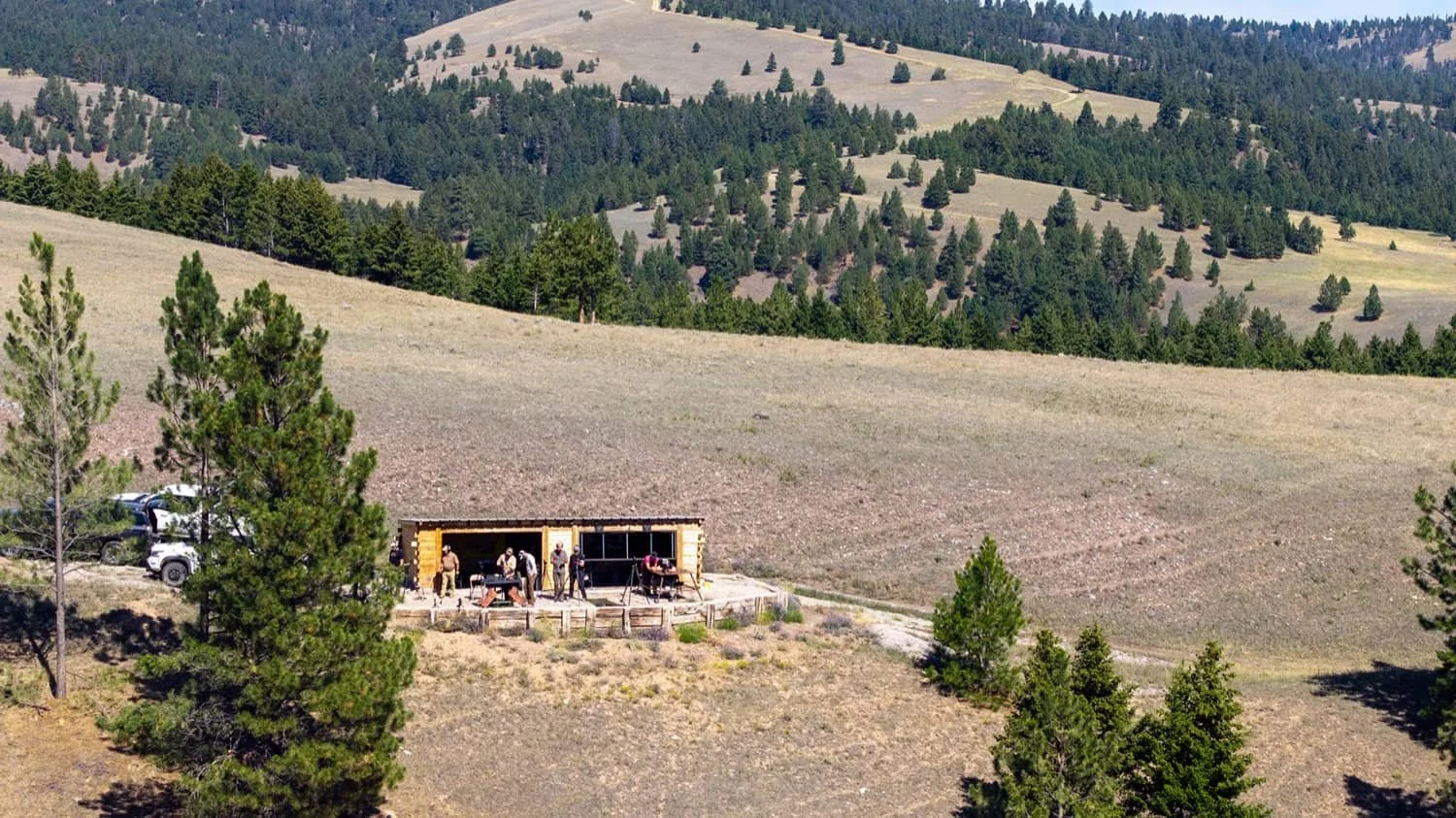 A hillside with a small wooden structure where people are gathered, surrounded by pine trees and a mountainous landscape in the background.
