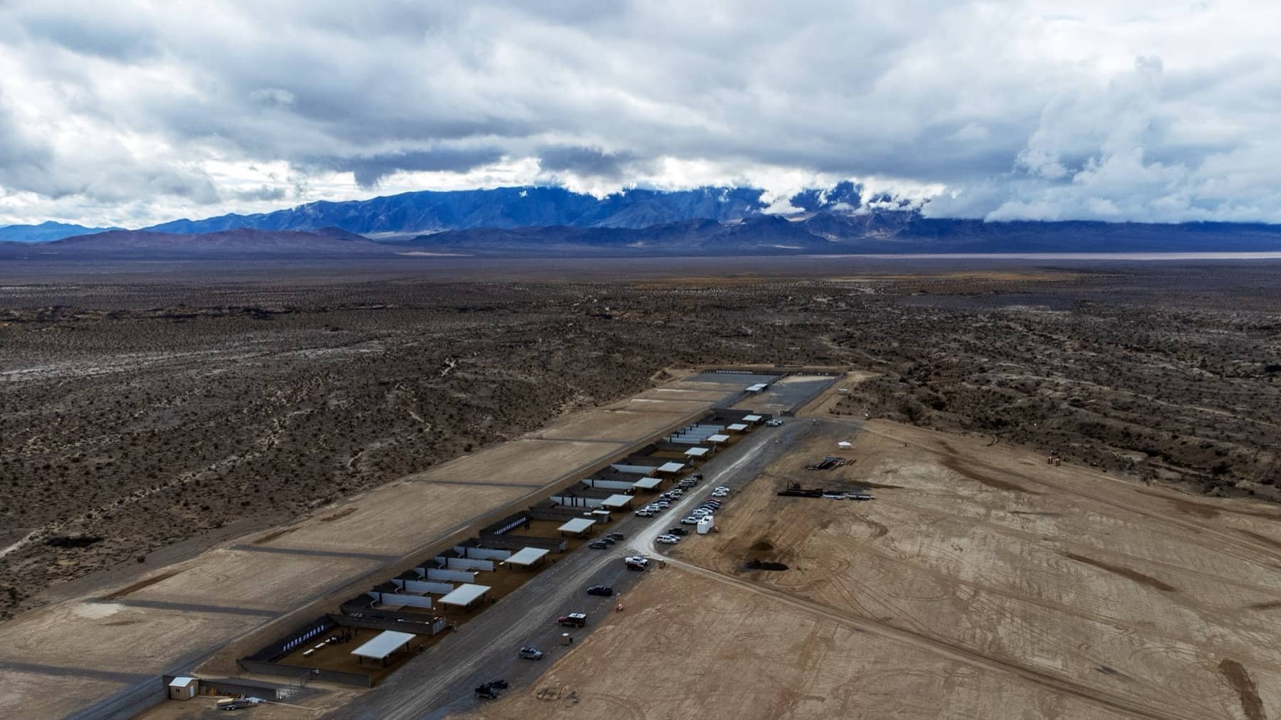 Aerial view of a border wall with multiple sections and security vehicles parked nearby in a desert landscape with mountains and cloudy sky in the background.