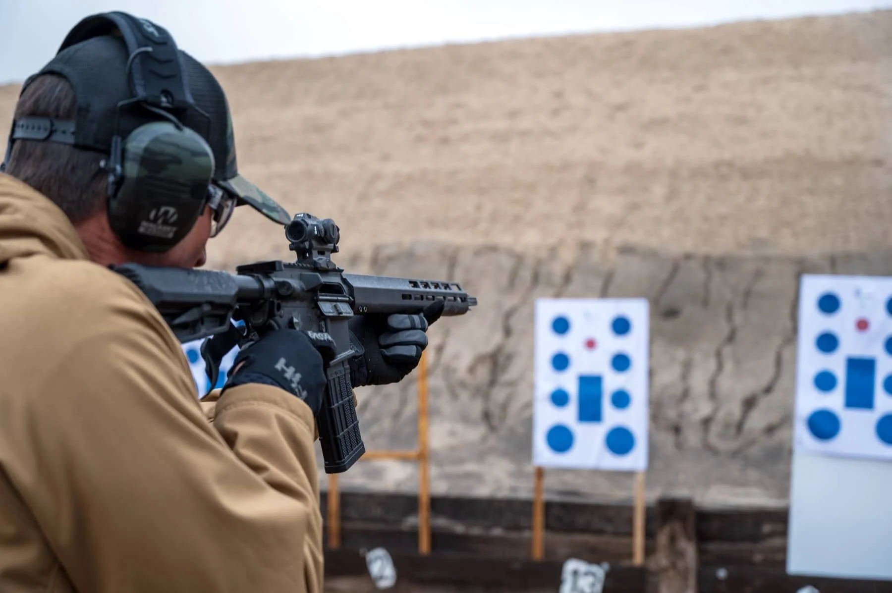 A man wearing a tan jacket, camouflage cap, and hearing protection, aiming a rifle at a shooting range with target boards in the background.