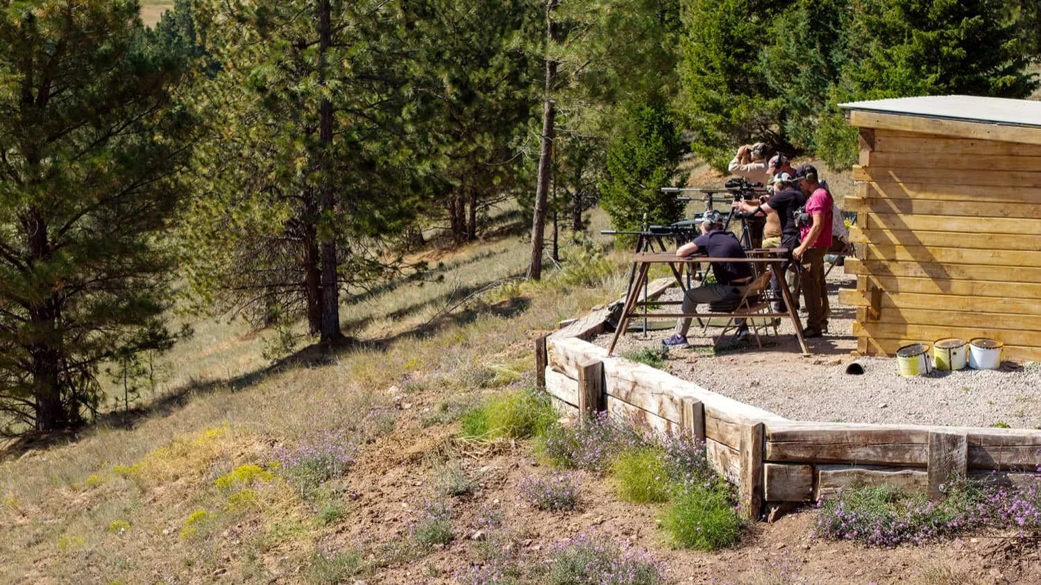 Group of people at an outdoor shooting range aiming rifles, surrounded by trees and natural landscape.