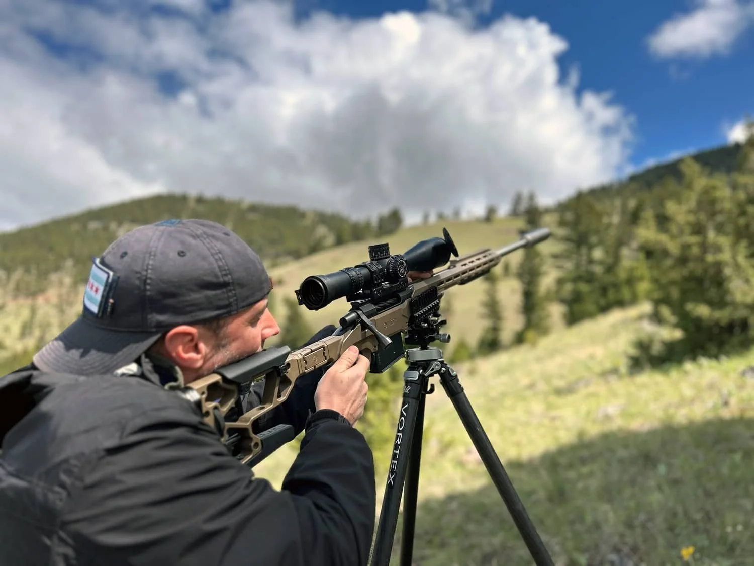 Man aiming a rifle with a scope outdoors on a tripod in a grassy mountainous area.