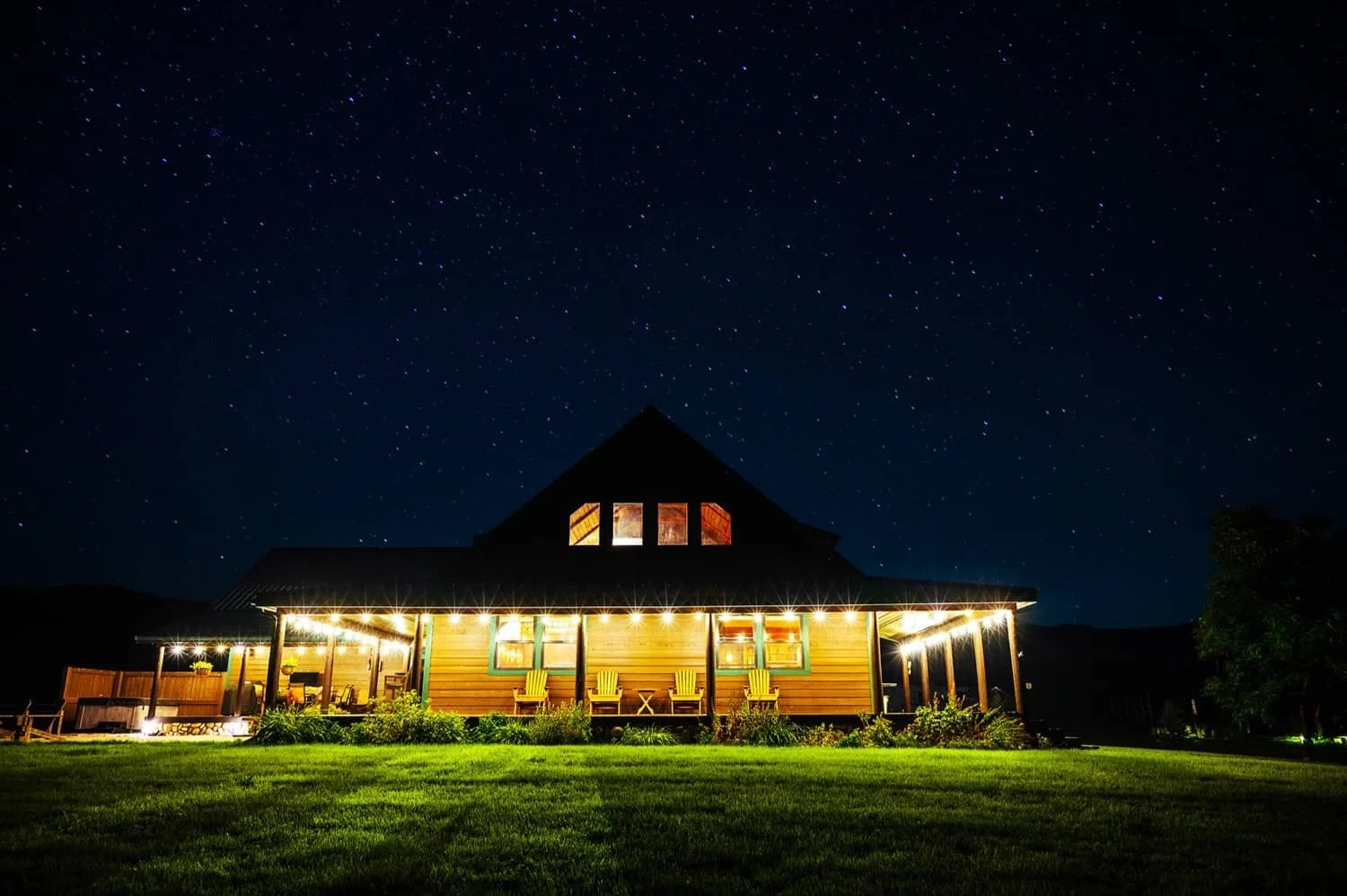 A cozy wooden cabin with a porch, illuminated with string lights, sits in a lush green yard under a starry night sky.