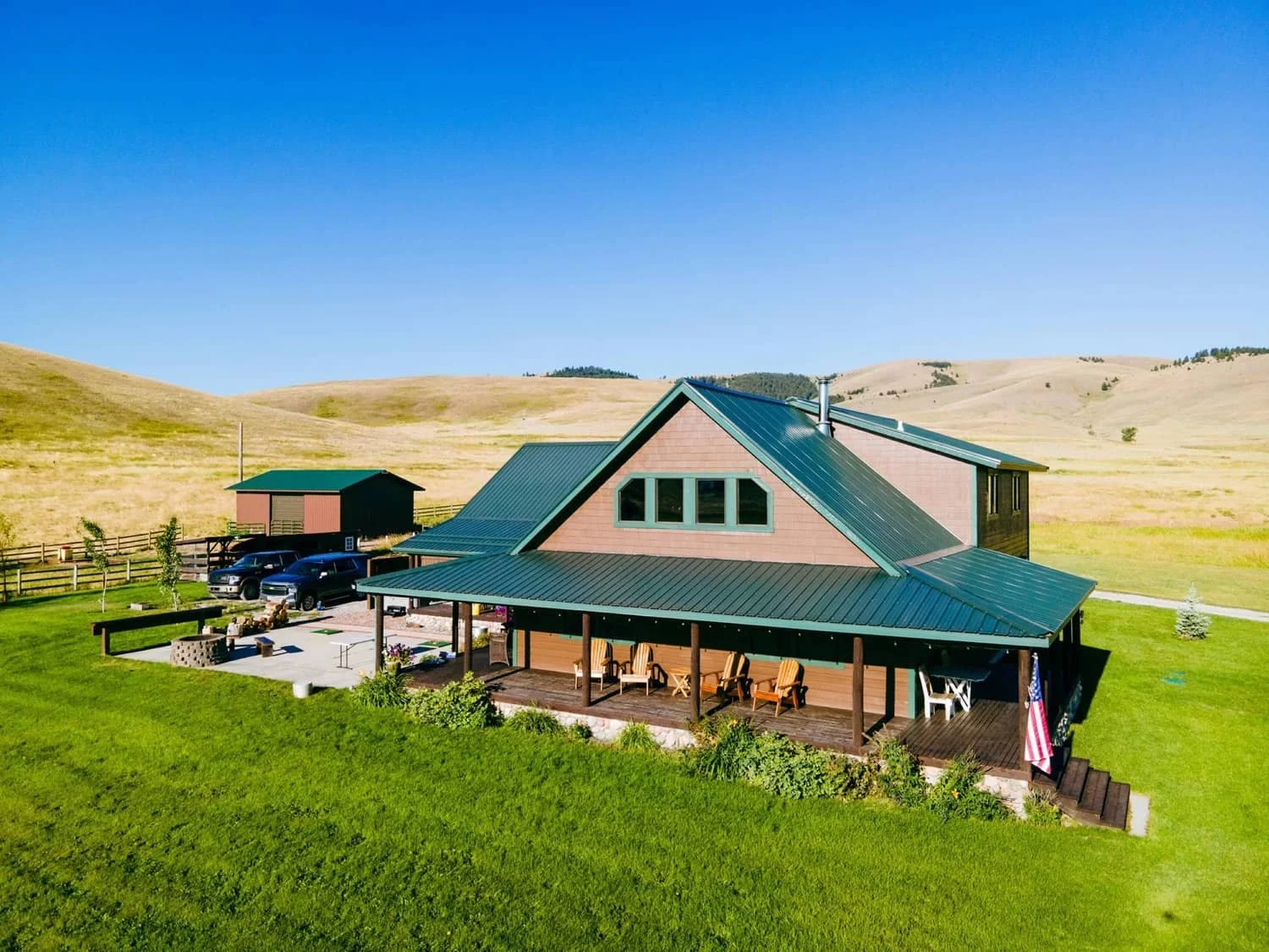 A countryside house with a green metal roof, surrounded by a vibrant green lawn and two small trees. The house has a large covered porch with outdoor furniture and an American flag. In the background, there are rolling hills and a clear blue sky.